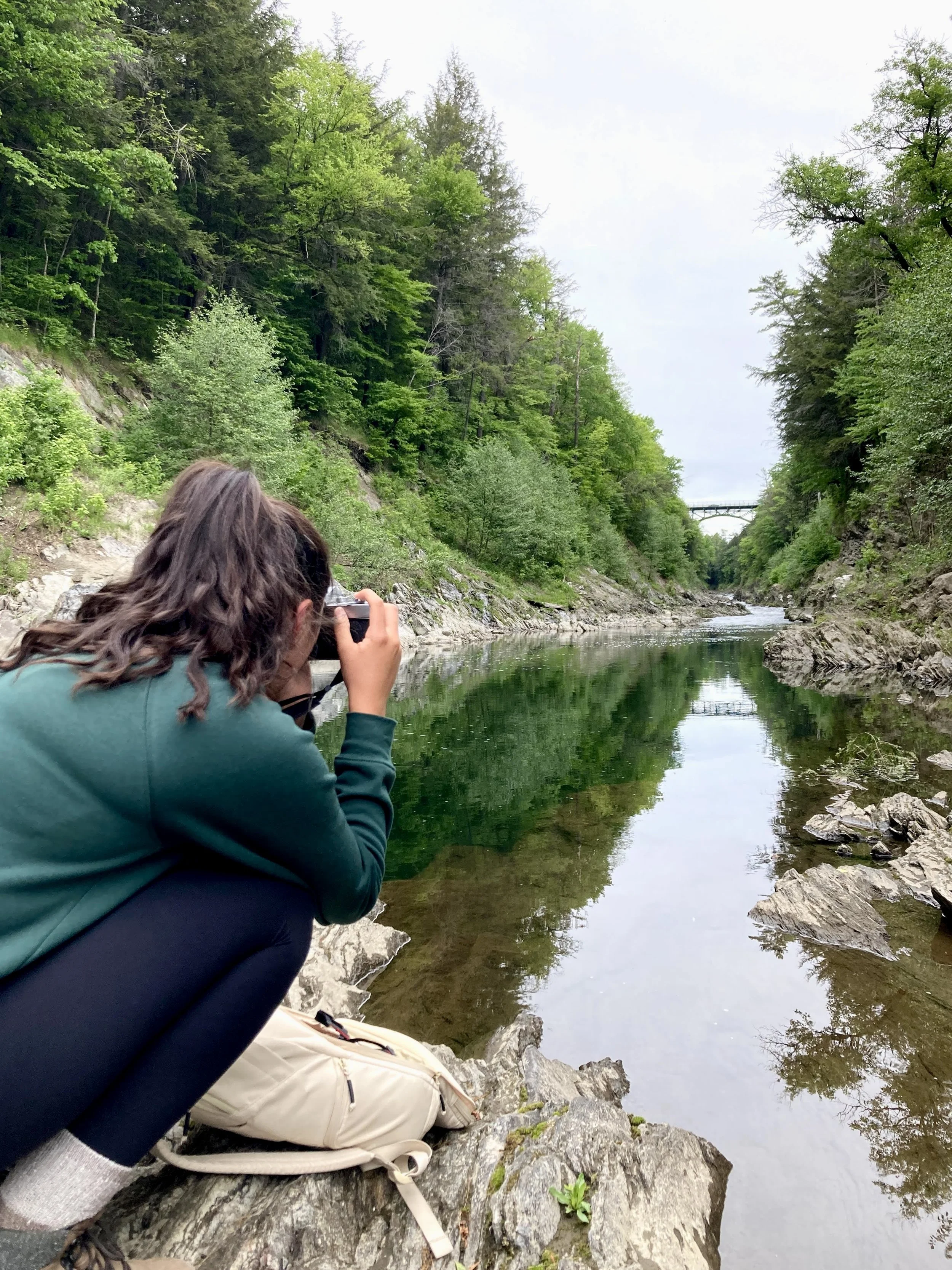 Person crouching on rocks taking a photograph of a calm river surrounded by green trees.