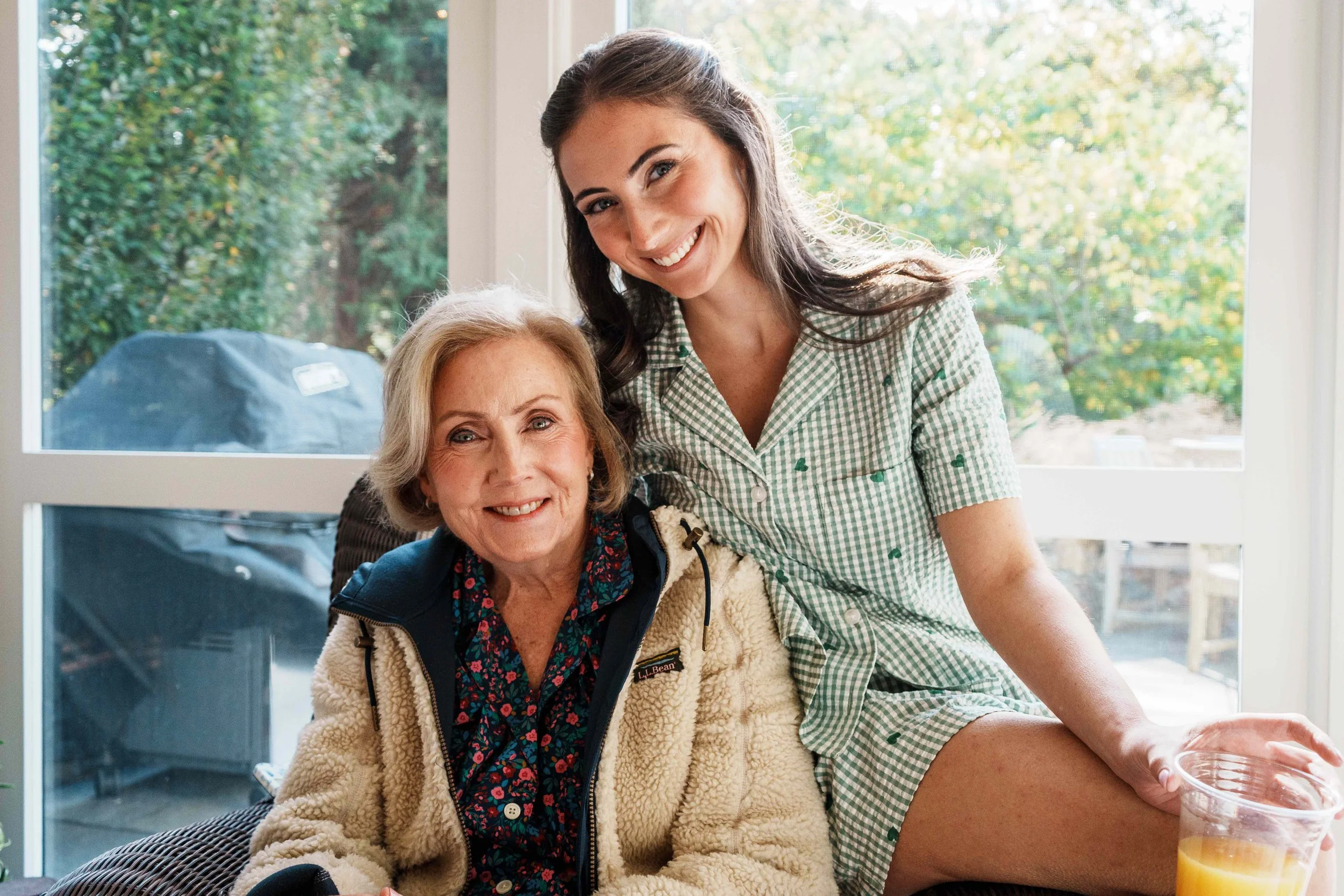 A young woman in green checkered pajamas sitting on the armrest of a chair with an elderly woman in a cream fleece jacket sitting beside her. They are smiling at the camera, and there is a large window with green trees outside in the background. The 