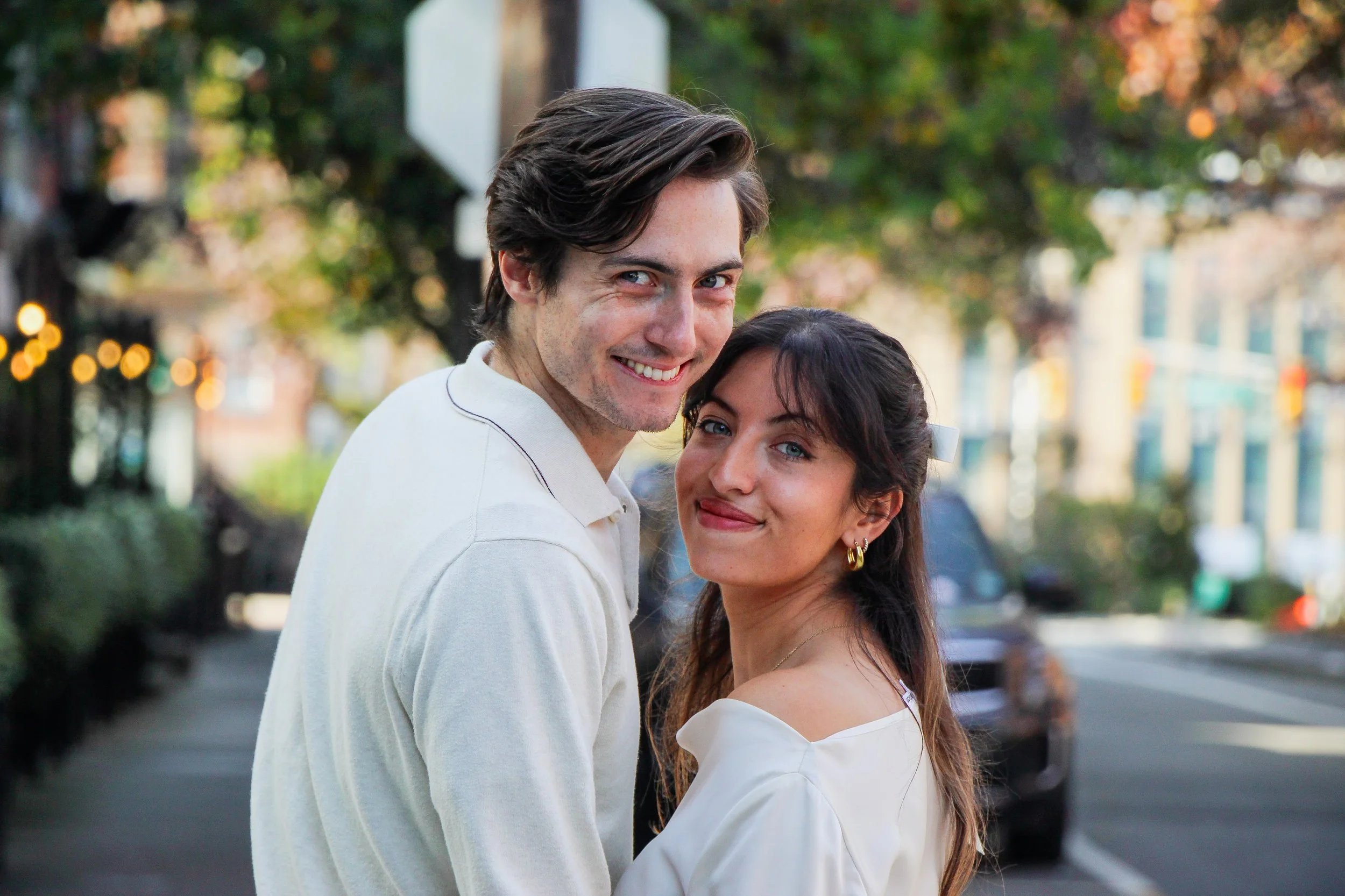 A smiling young couple standing close together outdoors in an urban setting with trees and parked cars, with the man wearing a white shirt and the woman wearing a white top and gold earrings.
