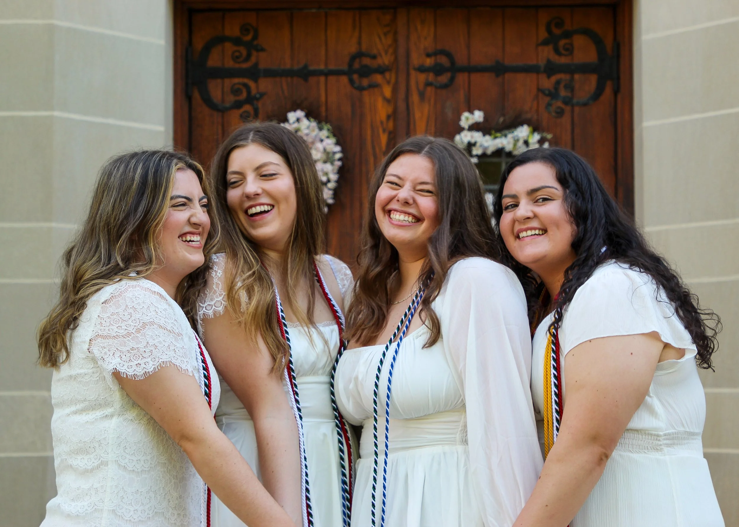 Four women in white dresses laughing and smiling together in front of a wooden door decorated with flower wreaths.