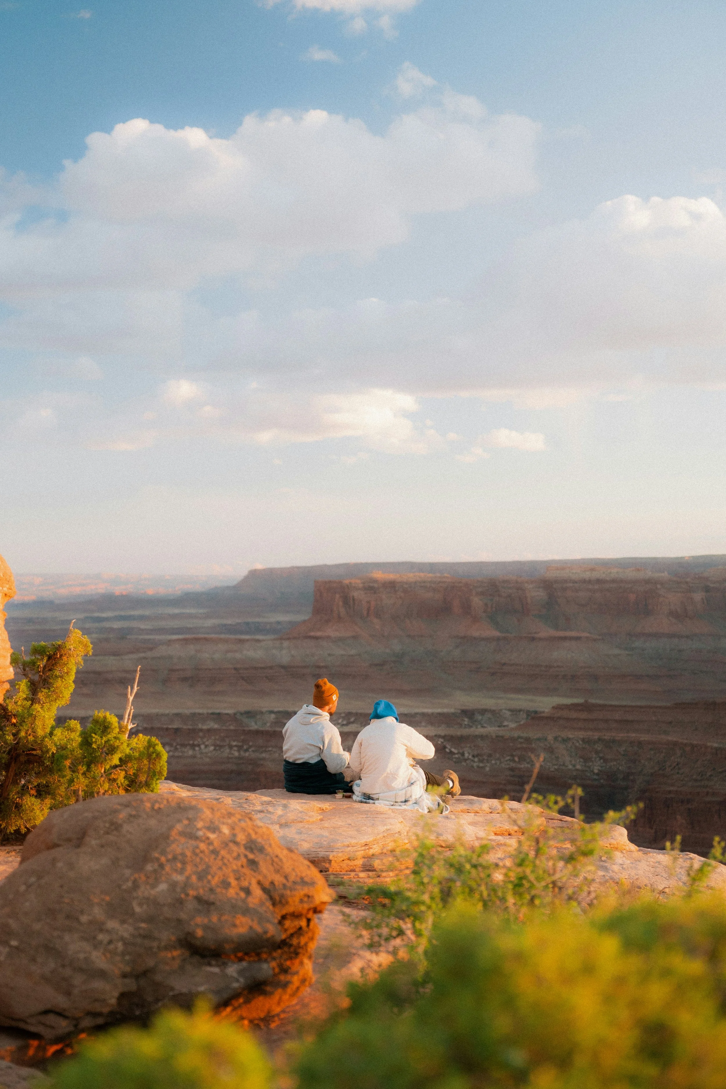 Two people sitting on a rocky ledge overlooking a canyon during sunset, with clouds in the sky.