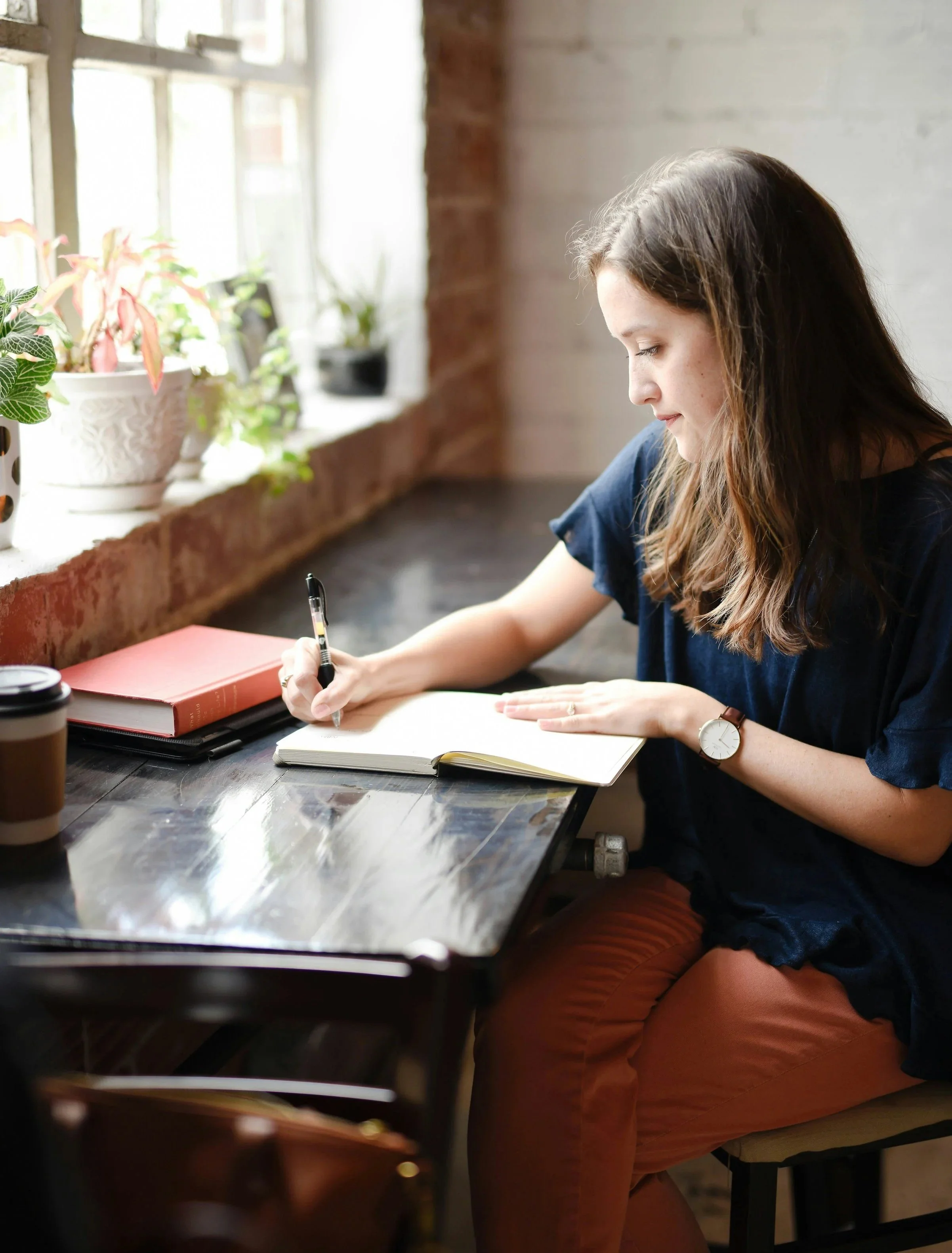 A woman sitting at a desk near a window, writing in a notebook with a pen, with books and a coffee cup on the desk, and potted plants on the windowsill.
