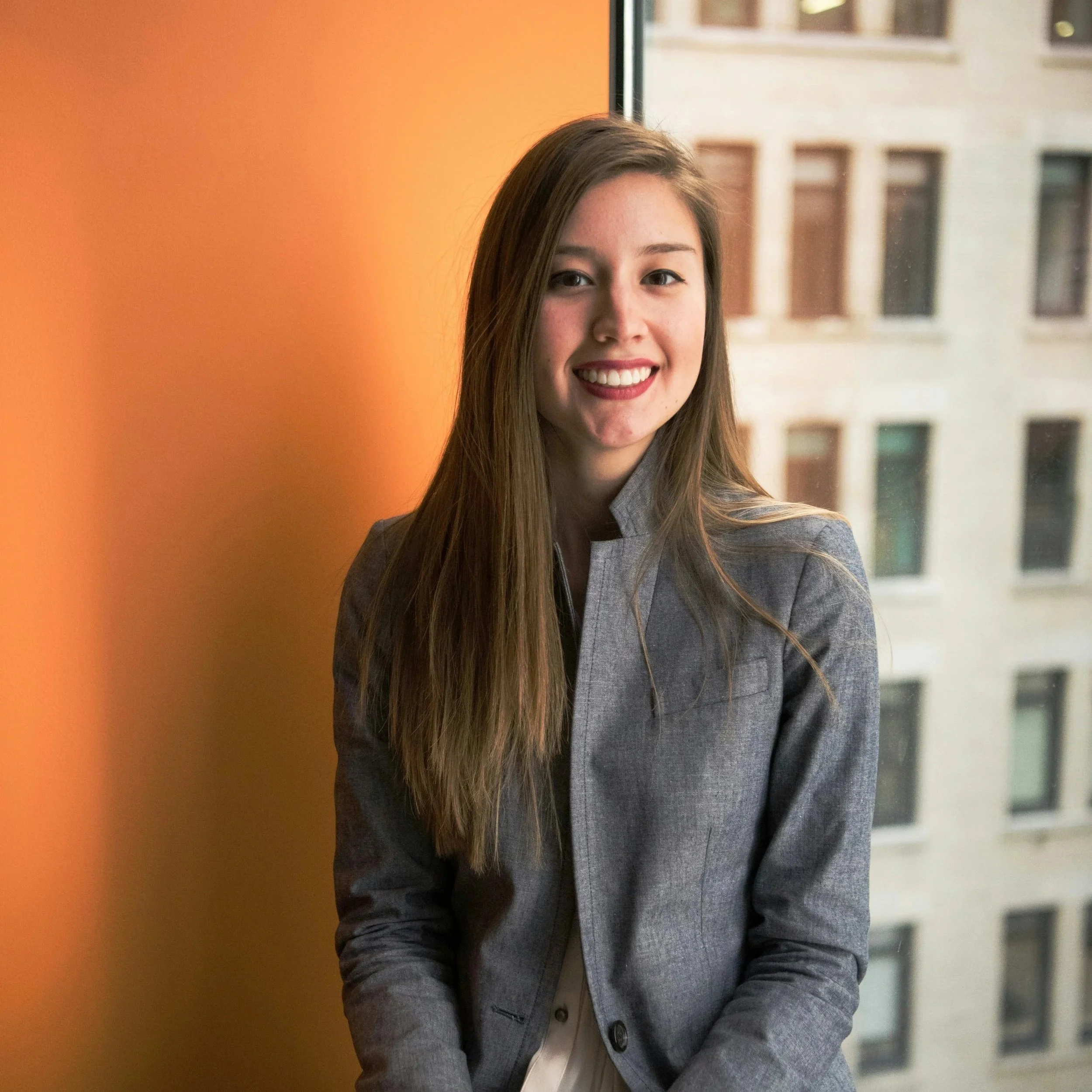 A young woman with long brown hair smiling, wearing a gray blazer and standing by a window with a cityscape view in the background.