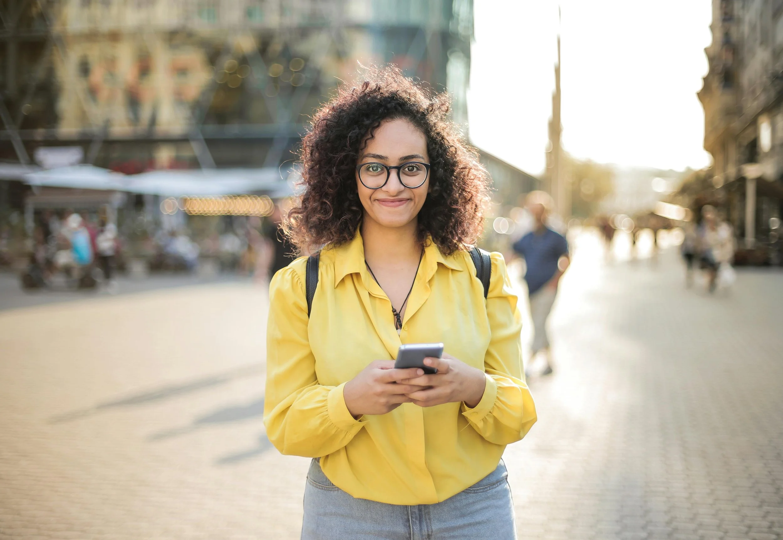 Young woman with curly hair, glasses, yellow blouse, and gray jeans standing on city street at sunset, looking at camera and holding smartphone.