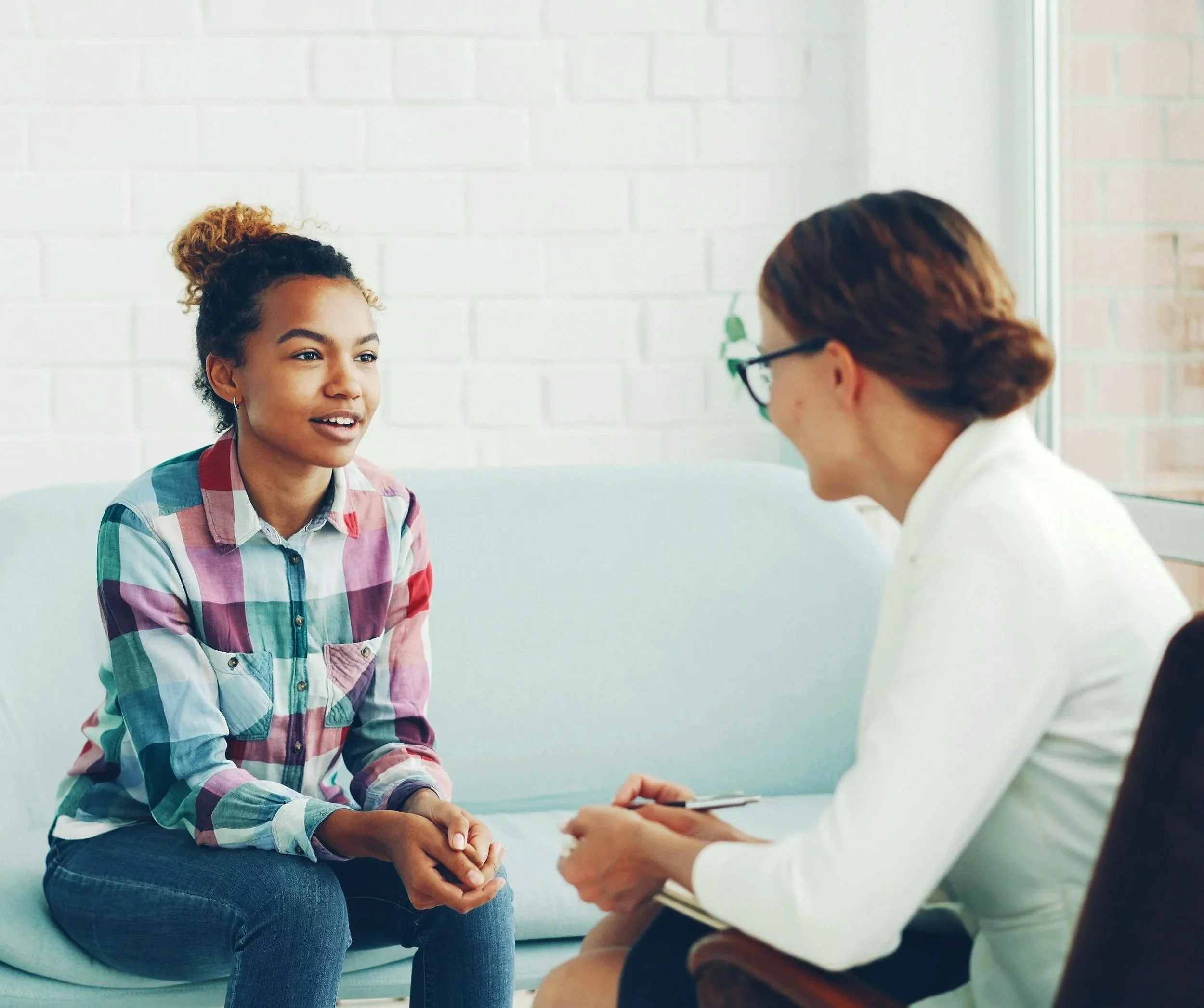 A young woman with curly hair, wearing a plaid shirt, sitting on a light-colored couch, and talking to a woman with glasses, wearing a white blazer, who is taking notes.