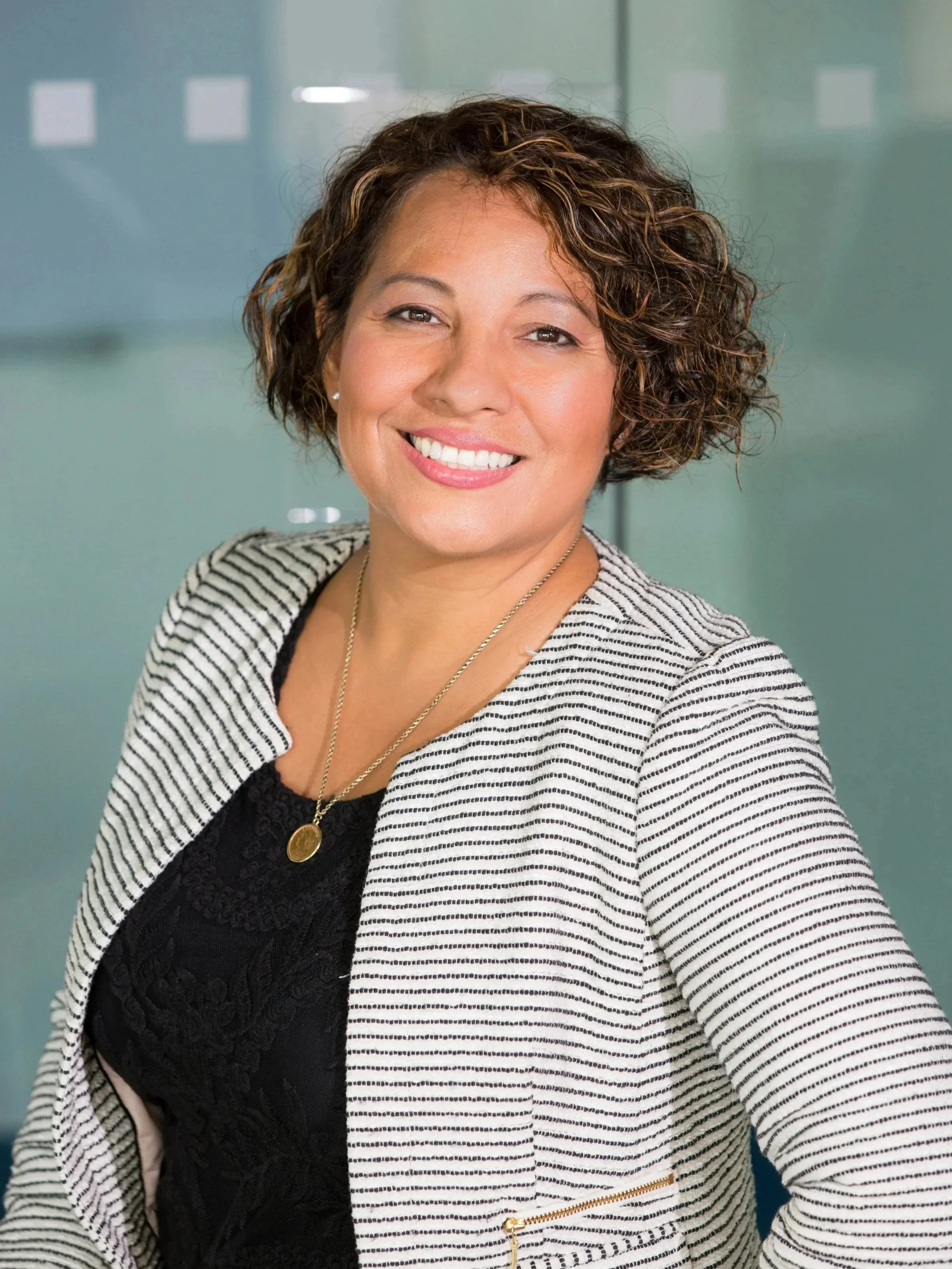 A smiling woman with curly, short brown hair in a business setting, wearing a striped blazer over a black lace top and a gold necklace.