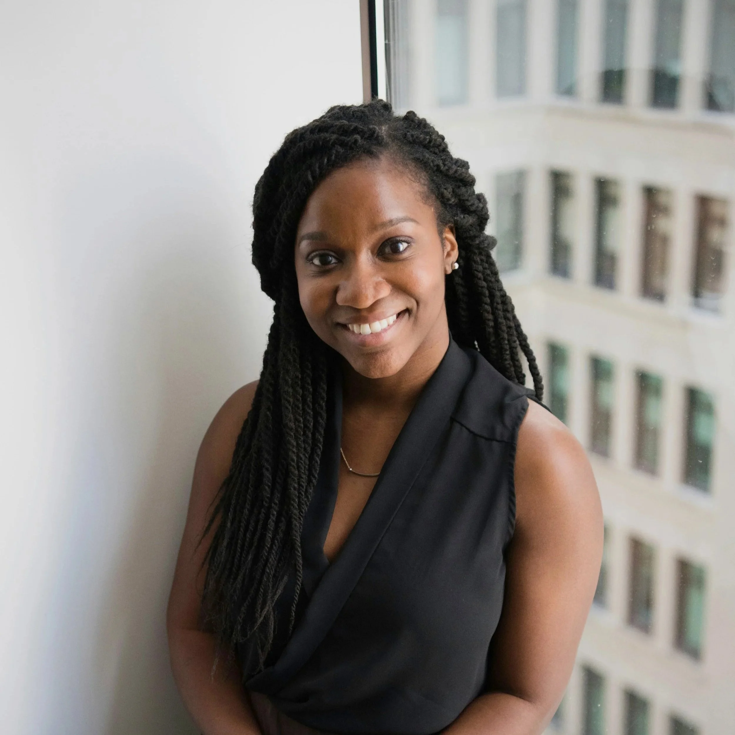 A woman with long, twisted braids wearing a sleeveless black top, smiling and standing next to a window in a high-rise building.