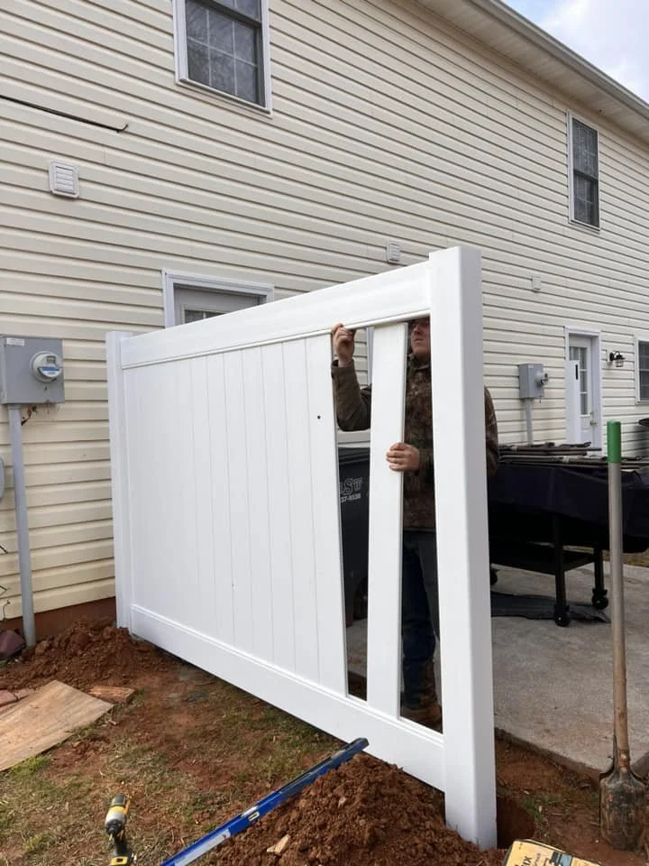 A person installing a white panel fence near a beige house with multiple windows, some tools and dirt in the yard.