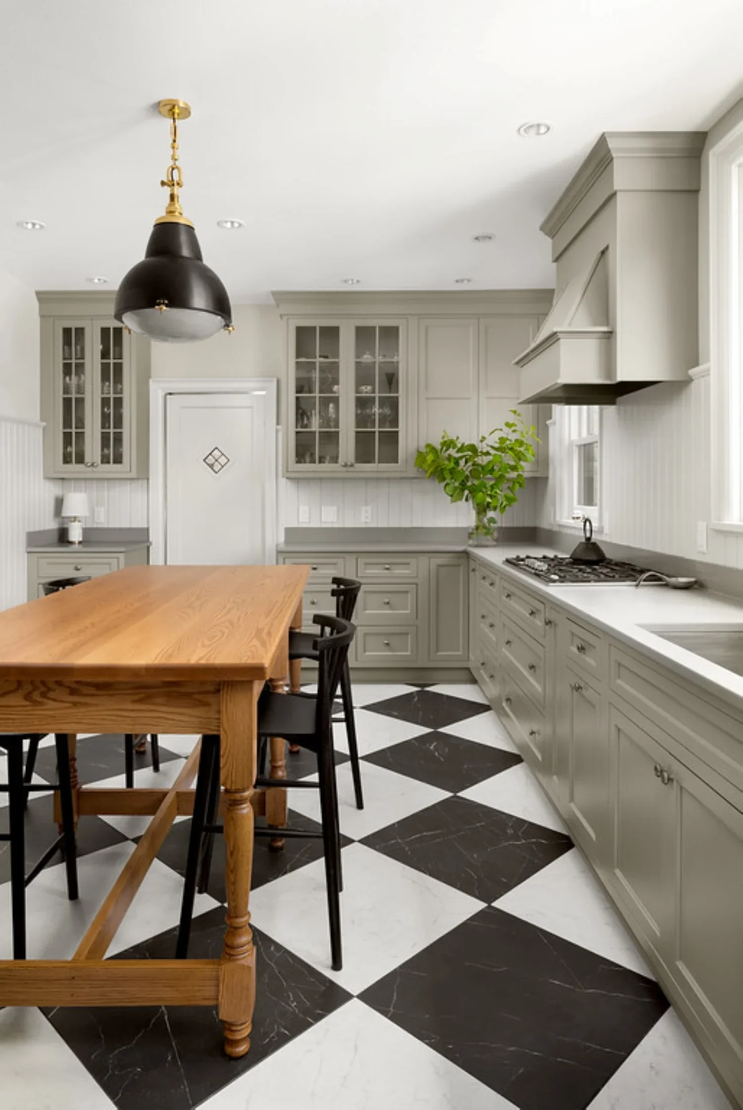 Modern kitchen with black and white checkered flooring, gray cabinets, wooden dining table, black chairs, and a black hanging light fixture.