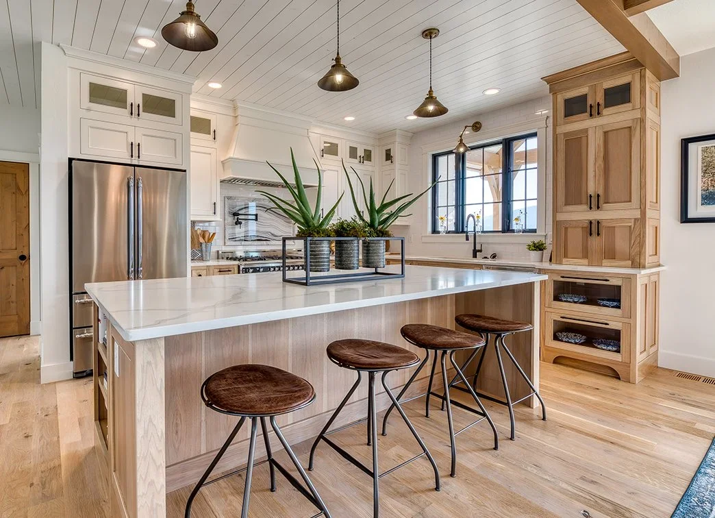Modern kitchen with white cabinets, stainless steel refrigerator, wooden accents, a large island with a white marble countertop, four brown bar stools, and potted plants on the island.