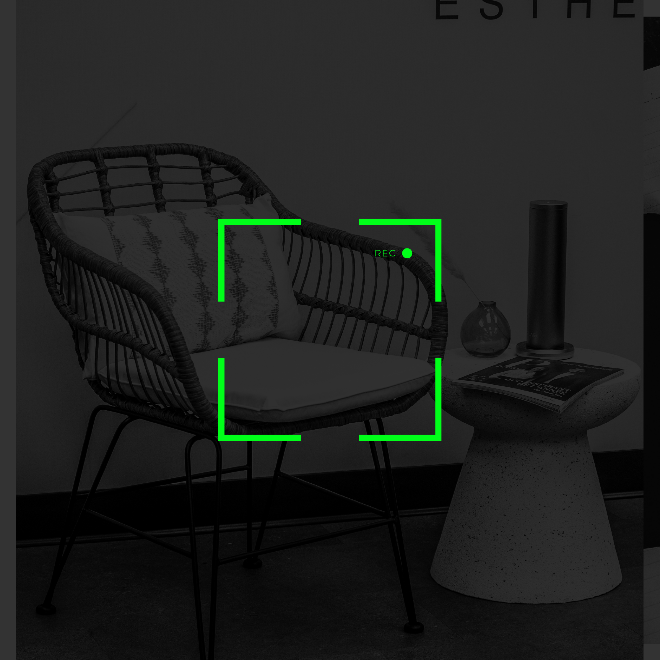 Un fauteuil en rotin avec coussin à rayures, une table en béton avec un magazine et deux vases en verre transparent, une lampe noire et un mur avec une affiche partielle.