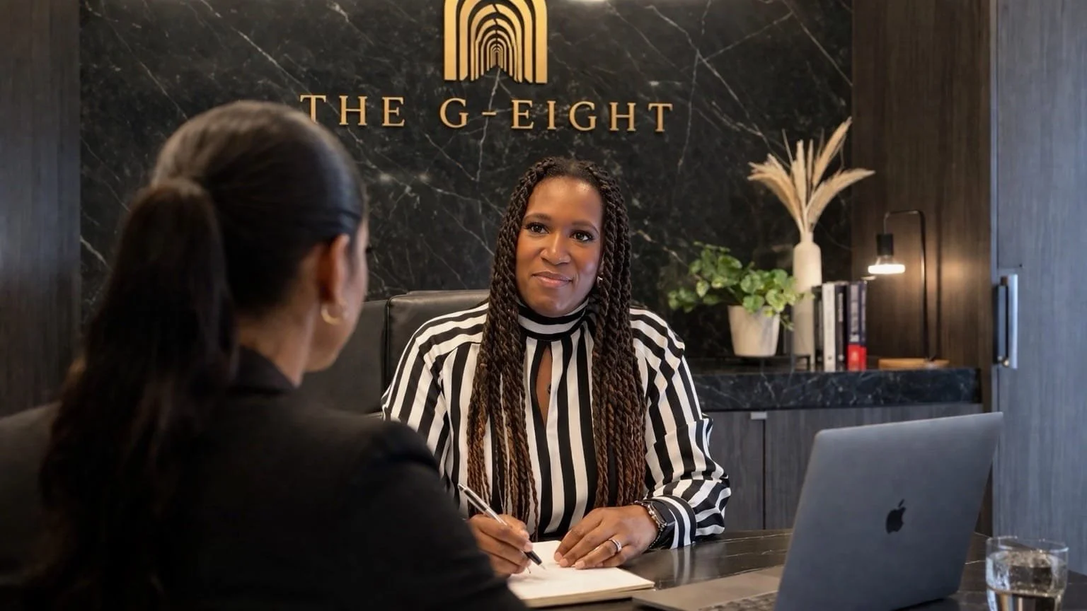 Two women are having a conversation in an office, seated at a desk with a laptop and a glass of water, in front of a black marble wall with gold lettering that reads "The G-Eight".