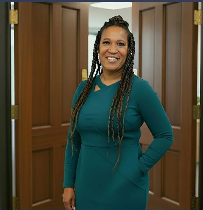 A smiling woman with long braided hair, wearing a teal dress, standing in front of wooden doors.