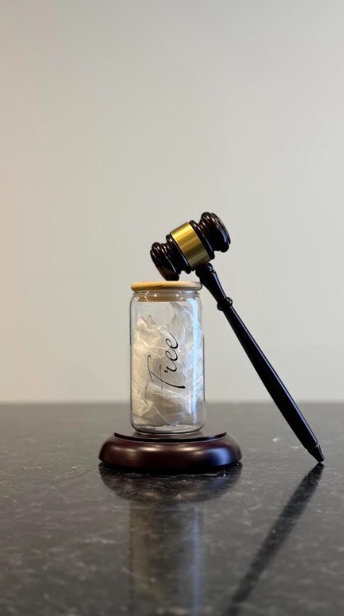 A glass jar labeled 'Feee' with crumpled paper inside, a gavel resting on top of the jar, and a microphone leaning against it. The items are on a dark countertop against a plain wall.