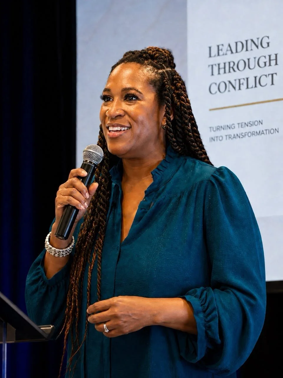 A woman speaking into a microphone at a conference, with a presentation slide titled 'Leading Through Conflict' in the background.