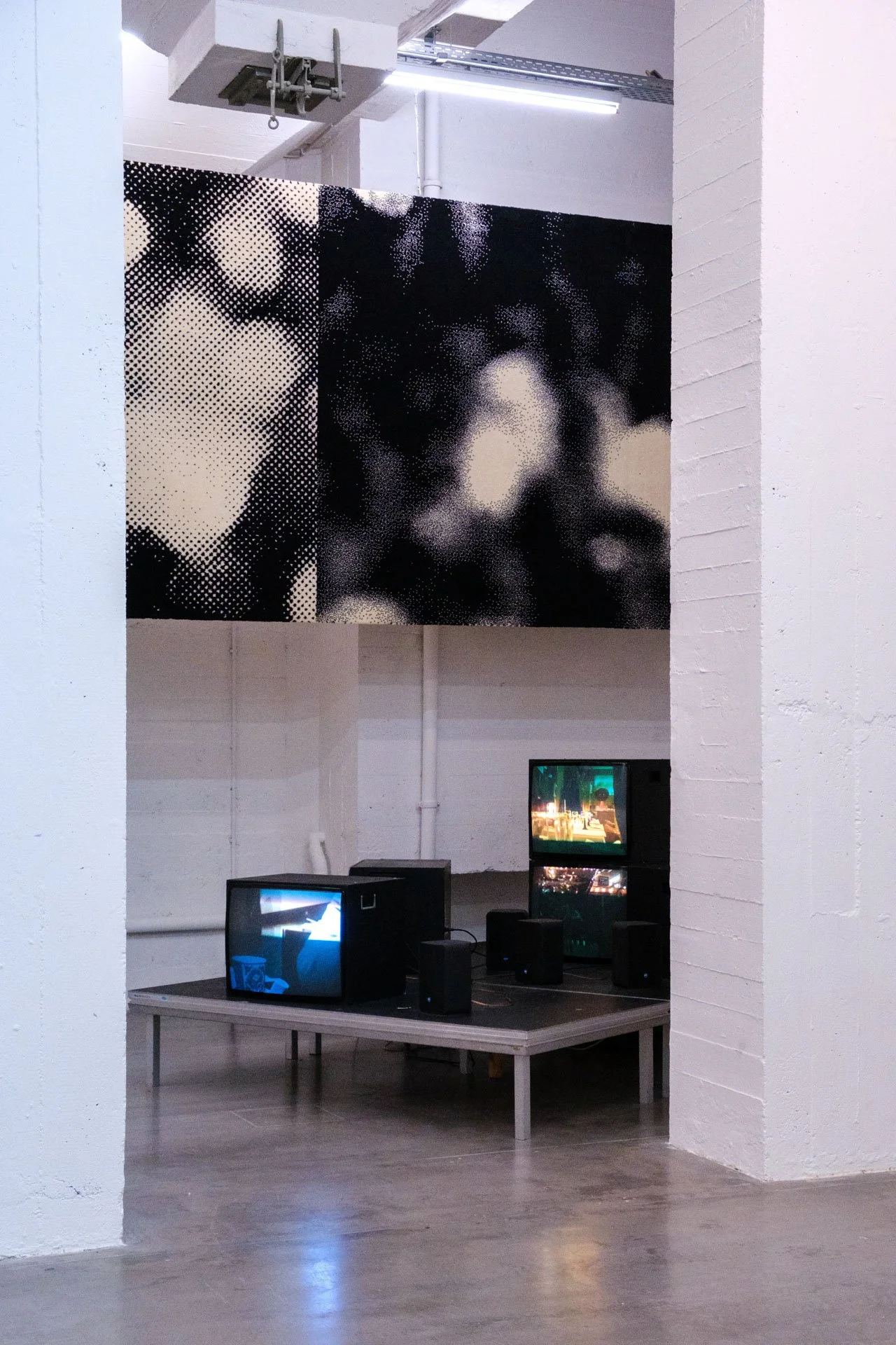 Installation artistique comprenant un panneau noir et blanc avec un motif de pointillisme représentant un visage, suspendu dans un espace blanc avec des murs en briques blanches, en dessous des ordinateurs portables avec écrans affichant différentes 