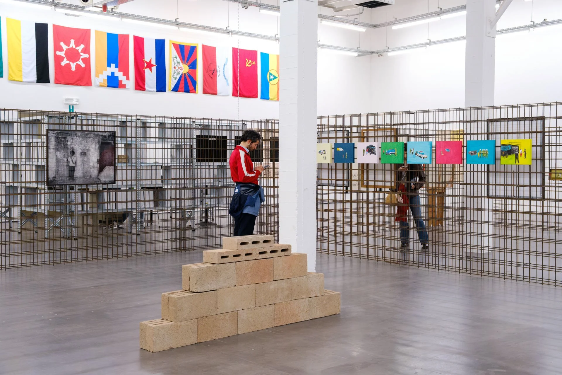 Une exposition d'art dans un espace neutre avec des drapeaux colorés accrochés au mur en haut. Au centre, un homme en rouge regarde son téléphone. Il y a des œuvres d'art ou des photographies accrochées sur une grille métallique, avec quelques person