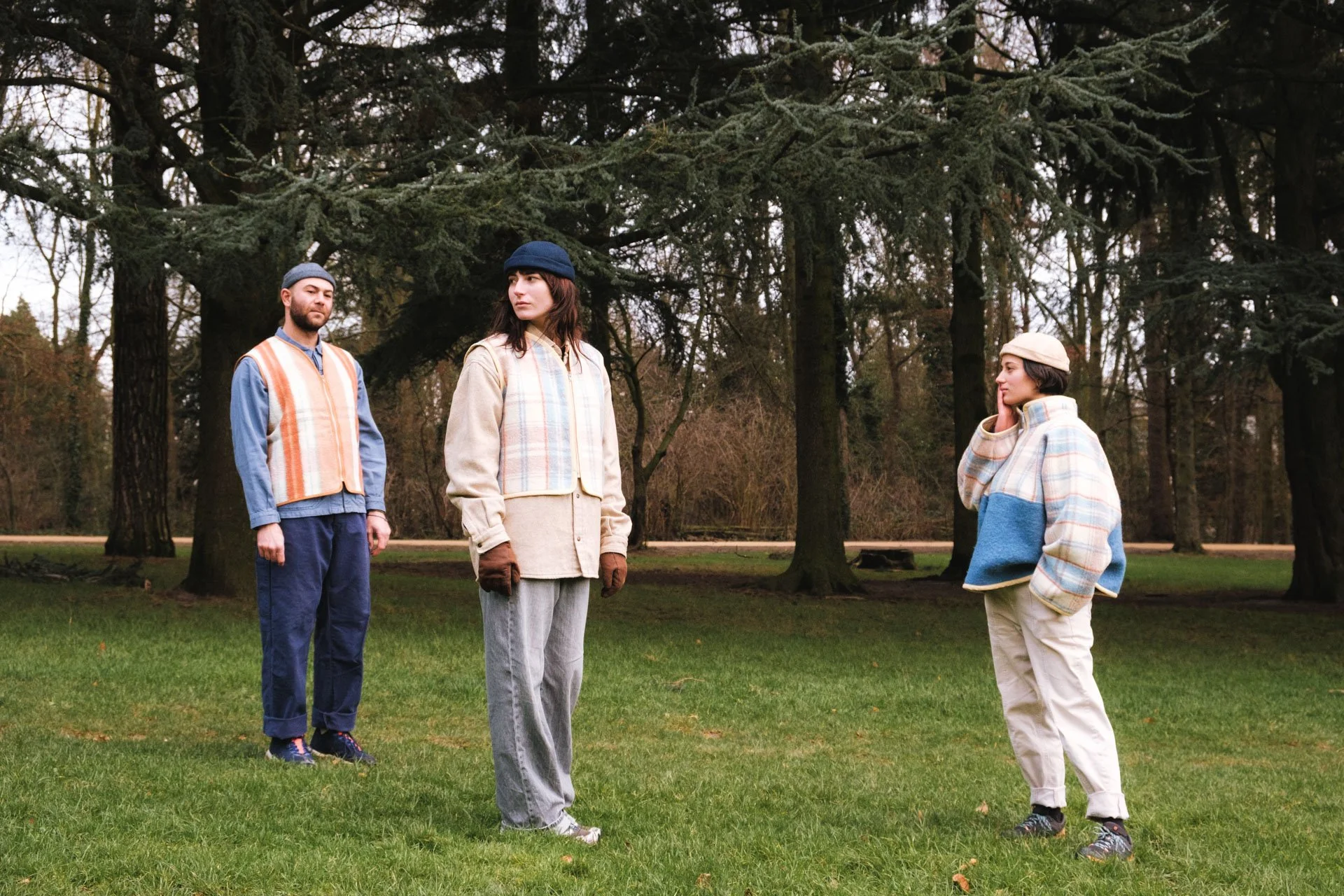 Trois personnes debout sur une pelouse dans un parc, avec des grands arbres en arrière-plan, portant des vêtements d'automne et regardant dans différentes directions.
