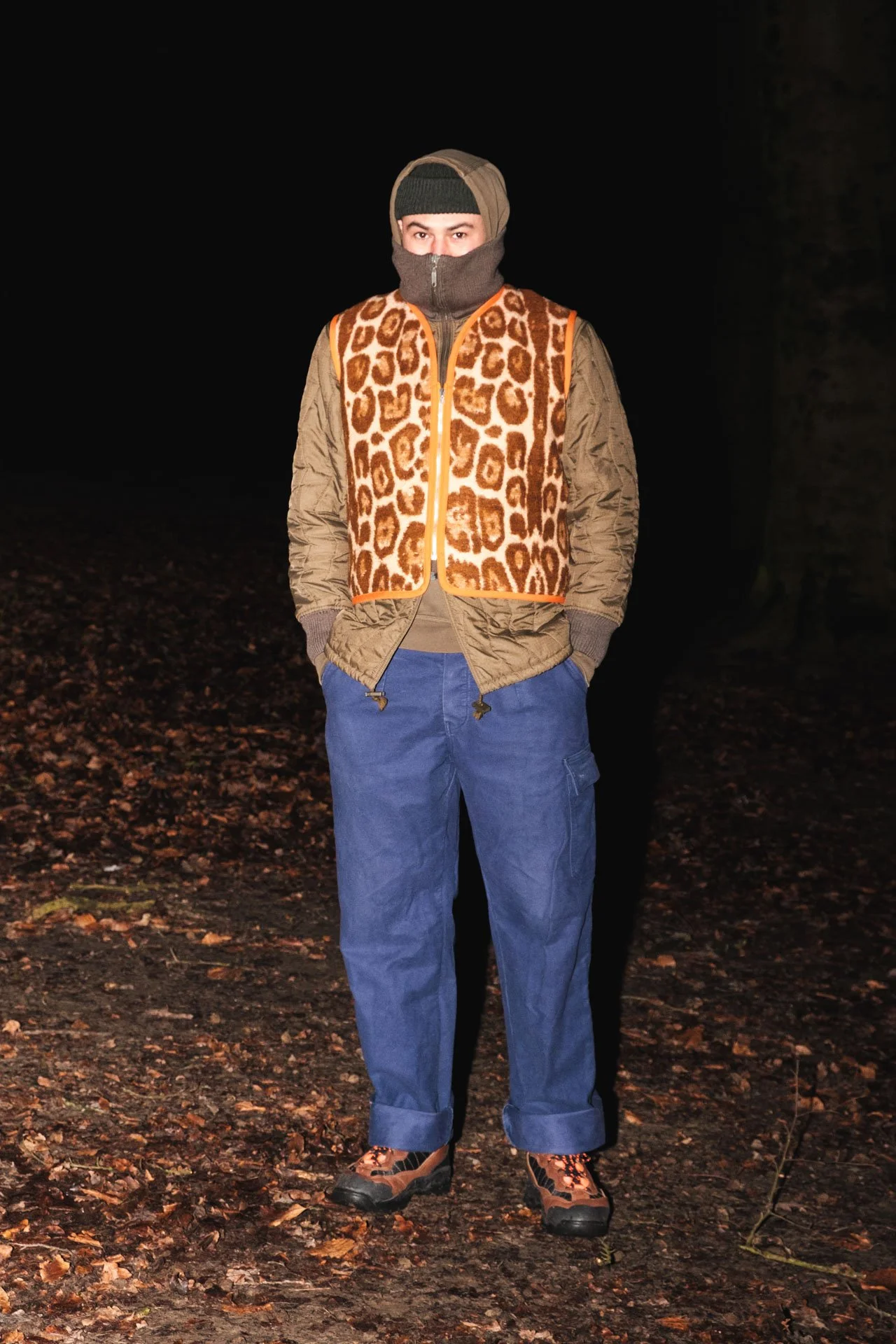 Une personne debout dans une forêt la nuit, portant des vêtements d'extérieur chauds, un pantalon bleu, une veste marron avec motif léopard orange, un bonnet et un cache-cou gris.