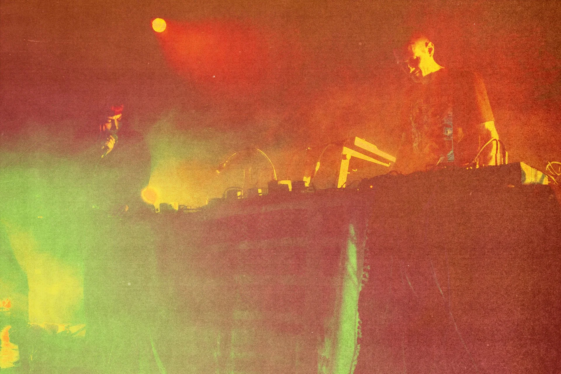 Deux hommes DJ en train de mélanger de la musique sur une scène éclairée par des lumières colorées orange et verte, avec un fond sombre.