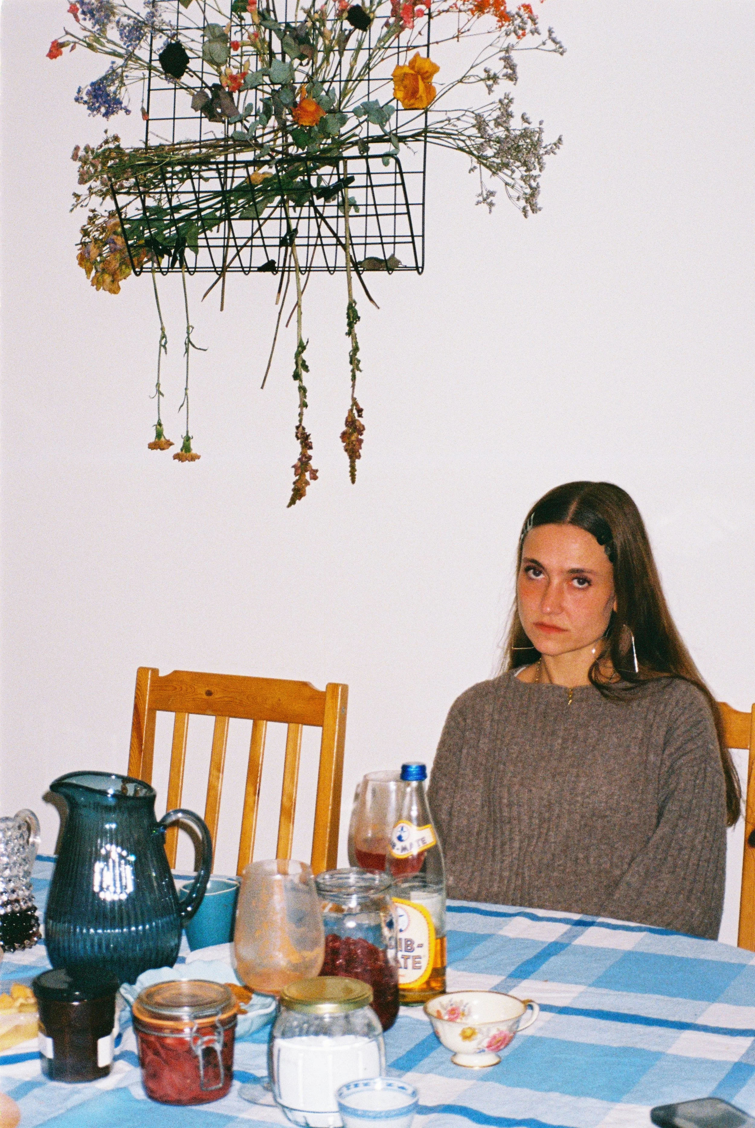 Une femme assise à une table avec des tasses, des bocaux de confiture, des plats et une bouteille d'eau. Au mur, un chandelier en fil de fer avec des fleurs sèches.