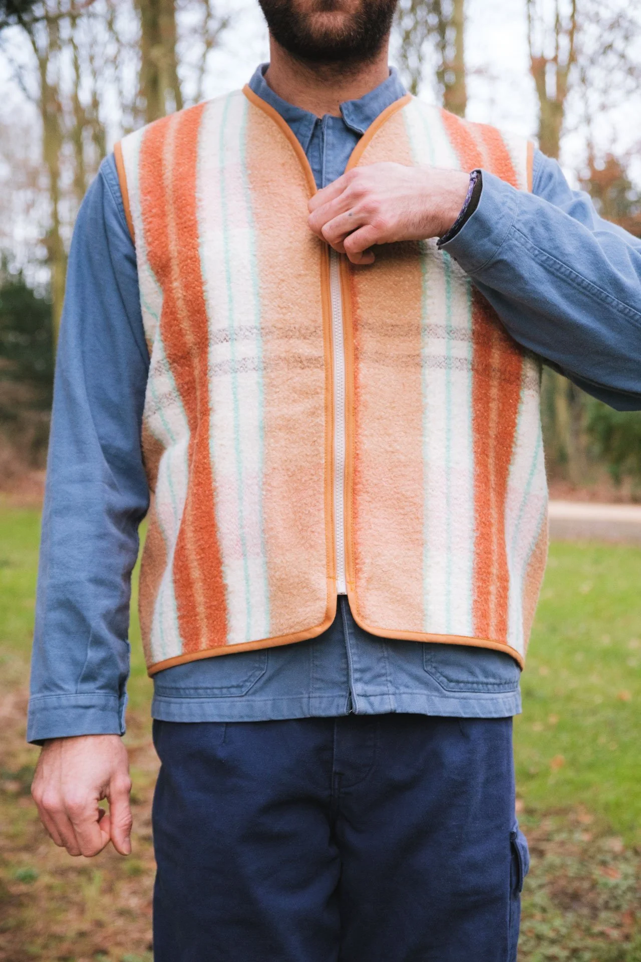 Homme portant un gilet sans manches orange à rayures et une chemise bleue, debout dans la forêt.