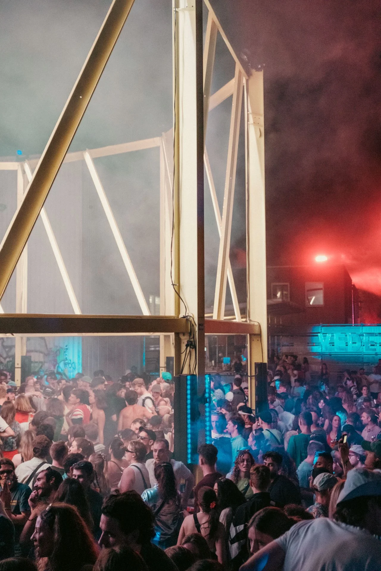 Une foule nombreuse assiste à un événement en soirée, avec une structure métallique en premier plan et des lumières colorées dans un environnement intérieur ou extérieur. Les gens discutent et semblent s'amuser.