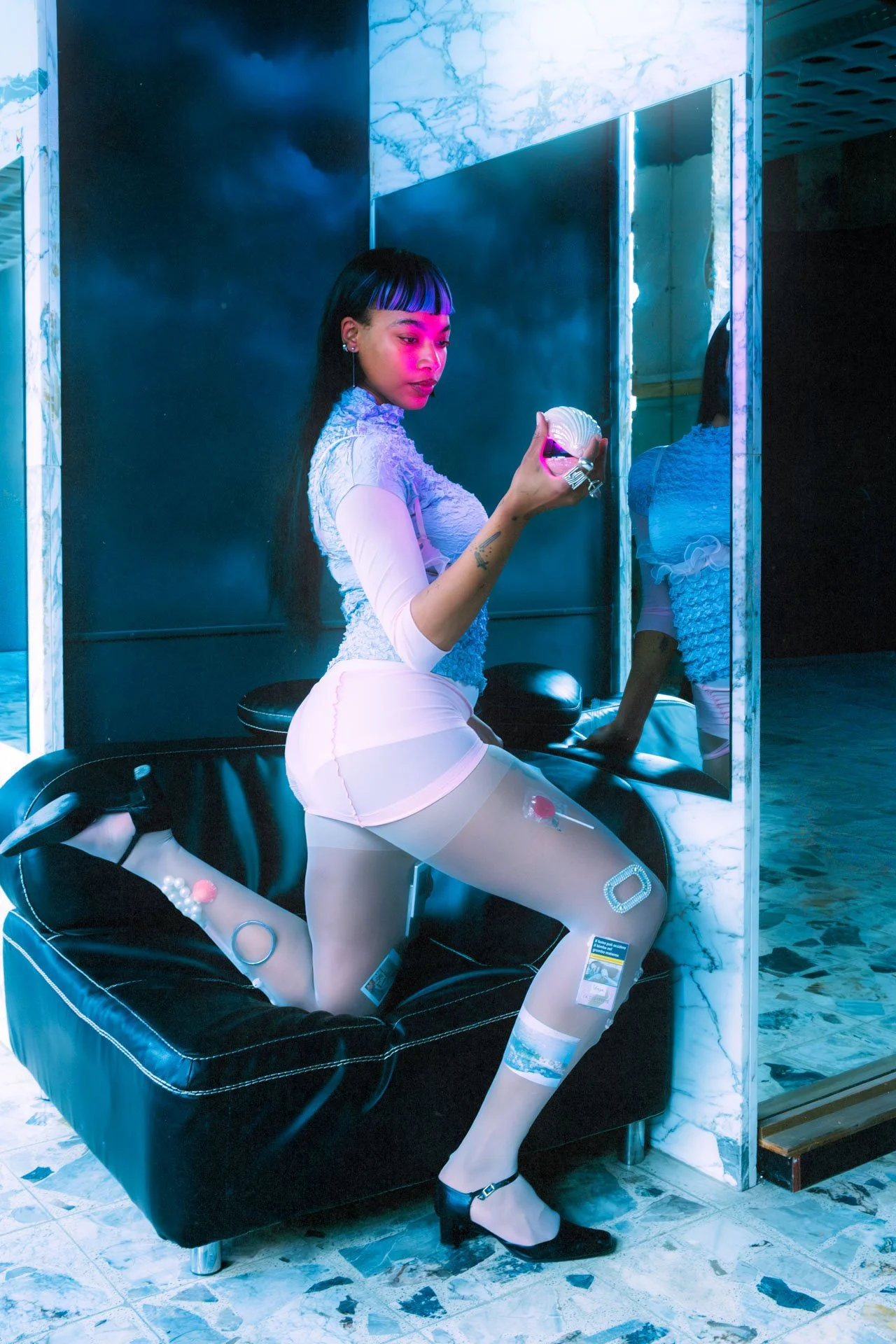 Une femme avec des vêtements futuristes et colorés posant sur un canapé noir, portant des collants transparents avec des motifs, devant une grande glace dans un intérieur moderne avec des lumières bleues et blanches.