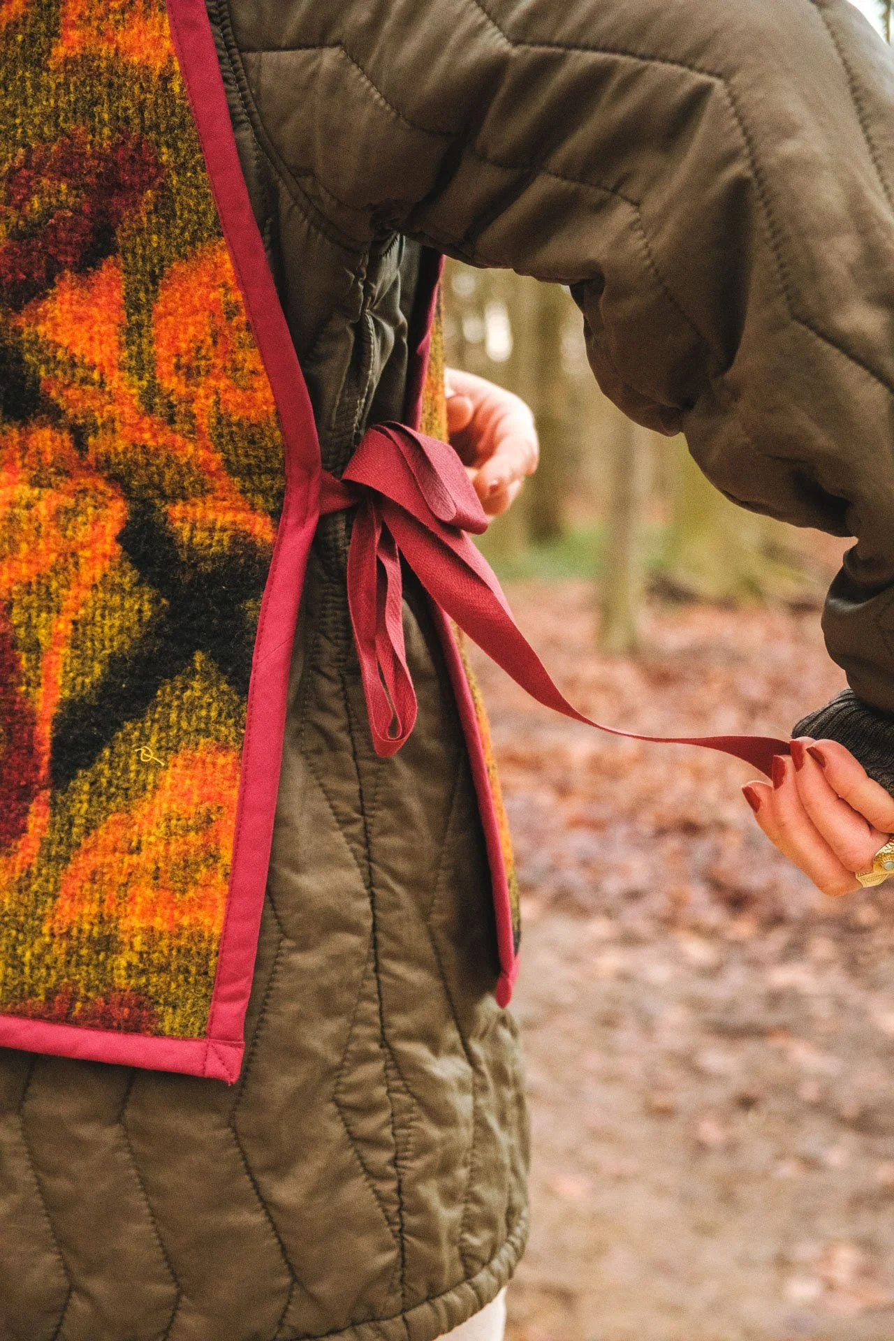 Une personne en pleine nature, portant une veste rembourrée beige et un foulard coloré avec des motifs automnaux, en train de faire un nœud avec un ruban rouge