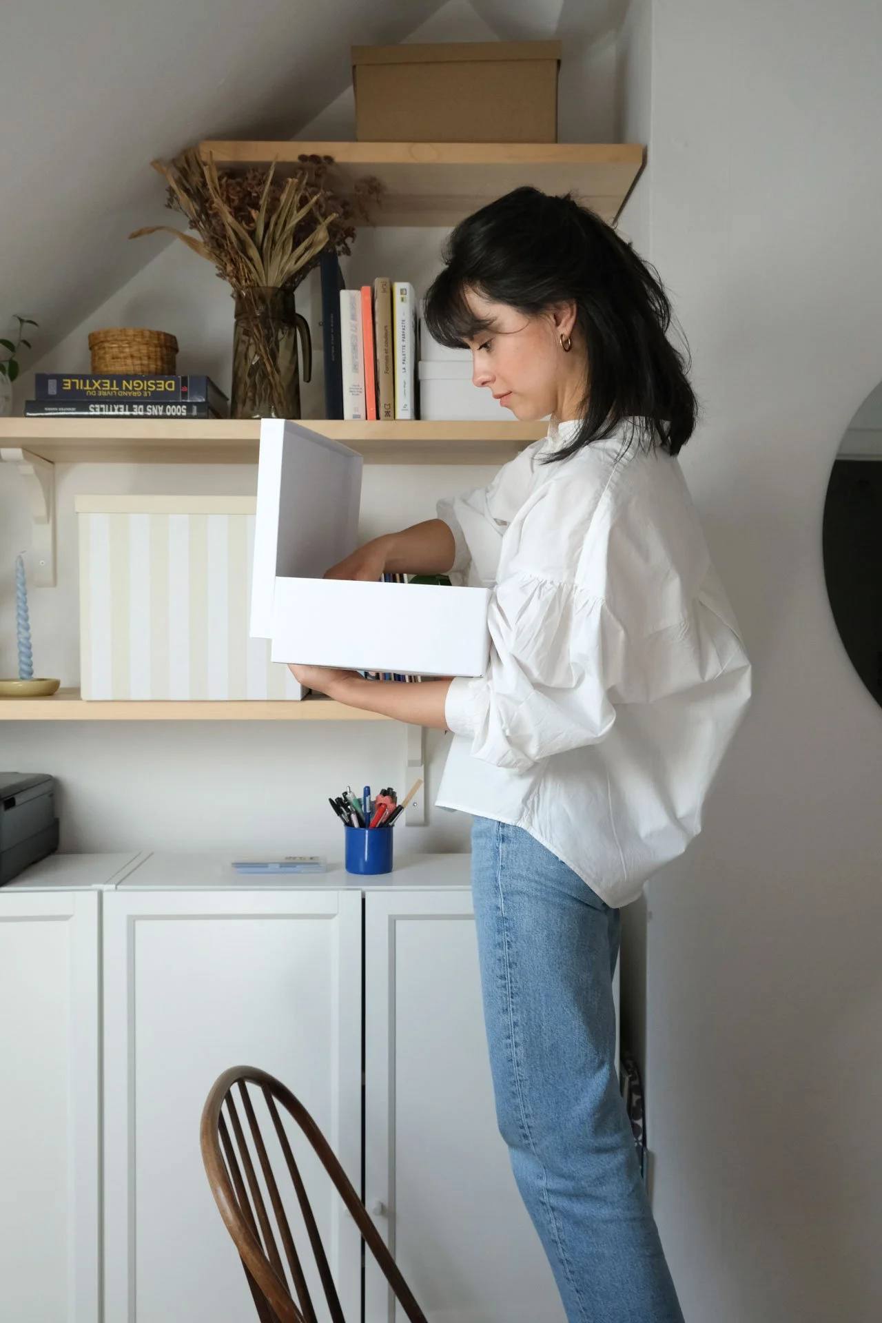 Une femme regarde dans une boîte blanche contenant des livres ou des documents dans un bureau à domicile, avec des étagères en bois et des objets décoratifs en arrière-plan.