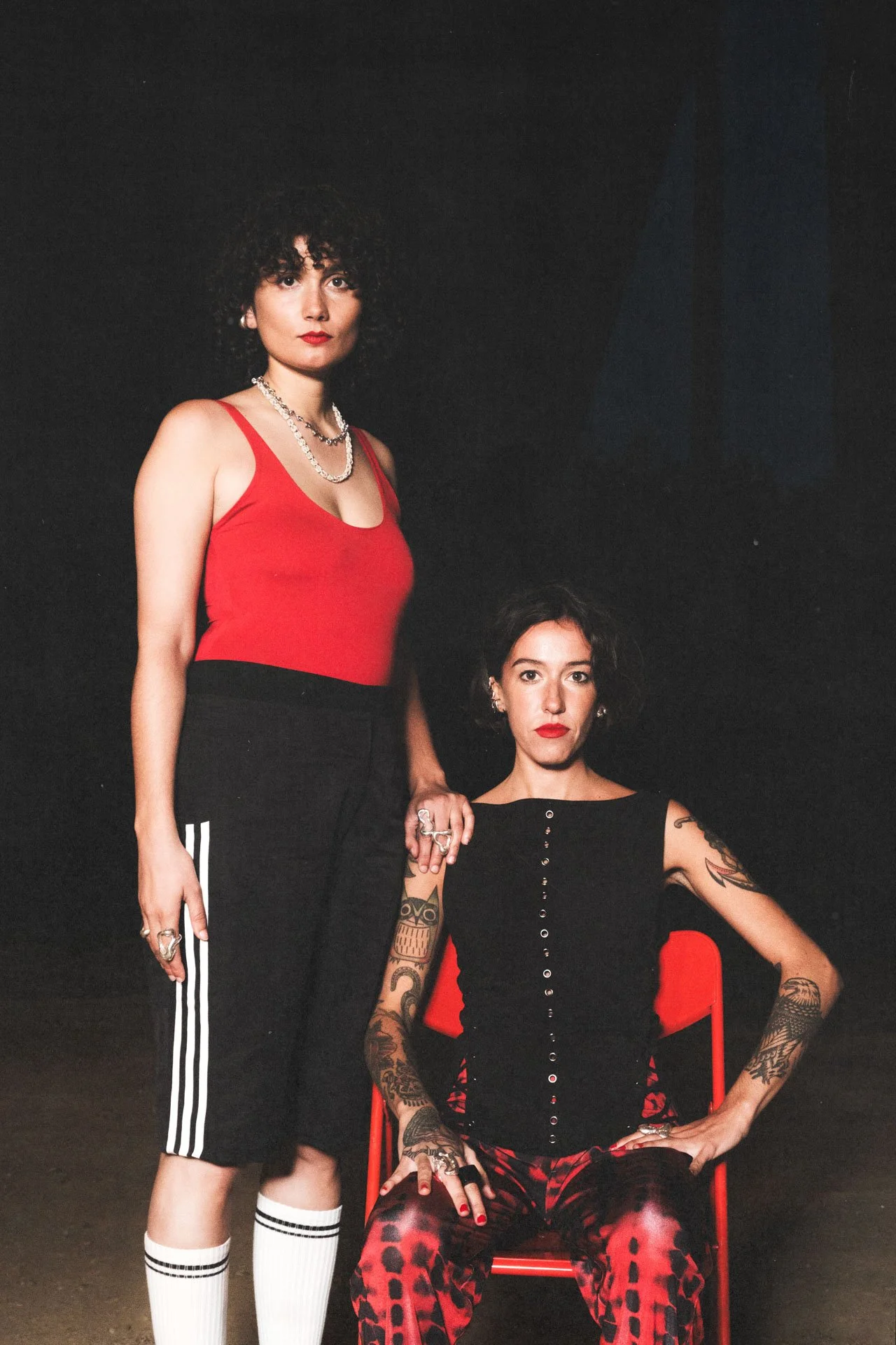 Deux femmes posant dans un studio sombre, l'une debout portant un débardeur rouge et un pantalon noir avec des rayures blanches, l'autre assise portant un haut noir ou rouge et un pantalon à motif rouge et noir, avec des tatouages visibles sur les br