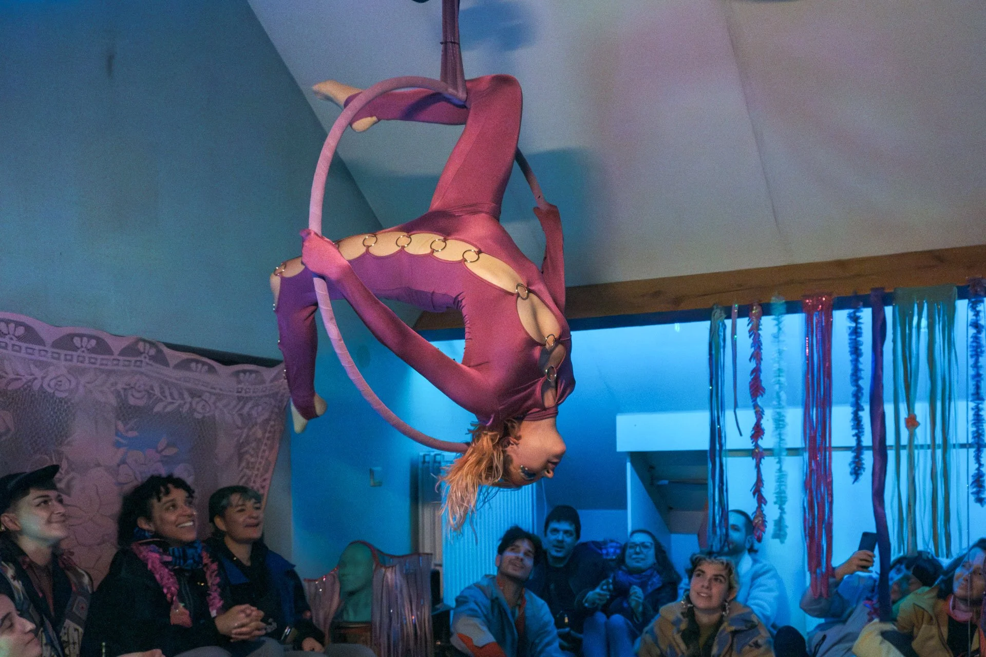 Une artiste réalise un numéro de cirque acrobatique dans une salle décorée de guirlandes colorées, avec un public d'admirateurs souriants qui l'observe.