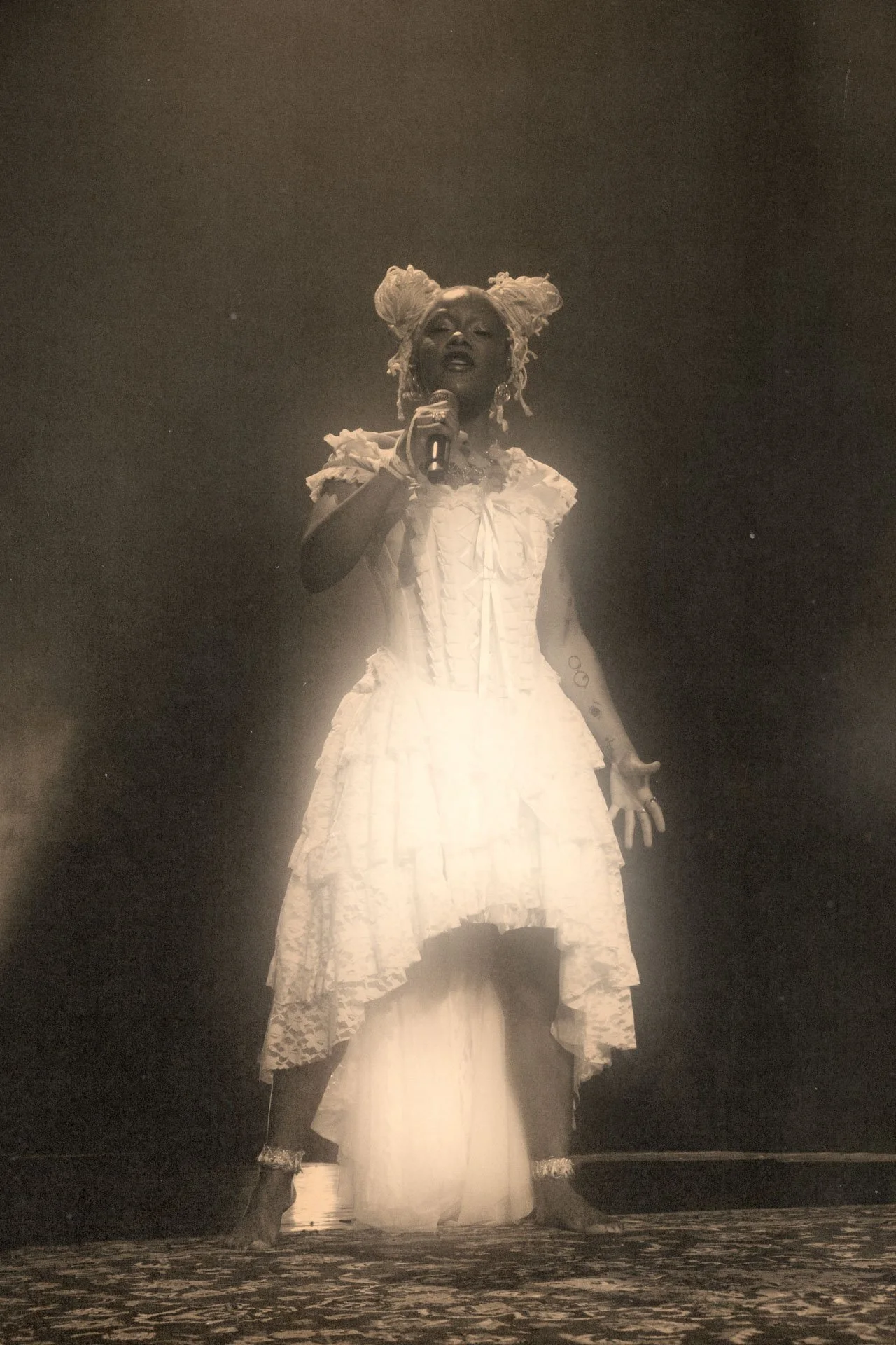 Une femme vêtue d'une robe blanche sophistiquée, tenant un microphone et se tenant sur une scène sombre avec un éclairage derrière elle.