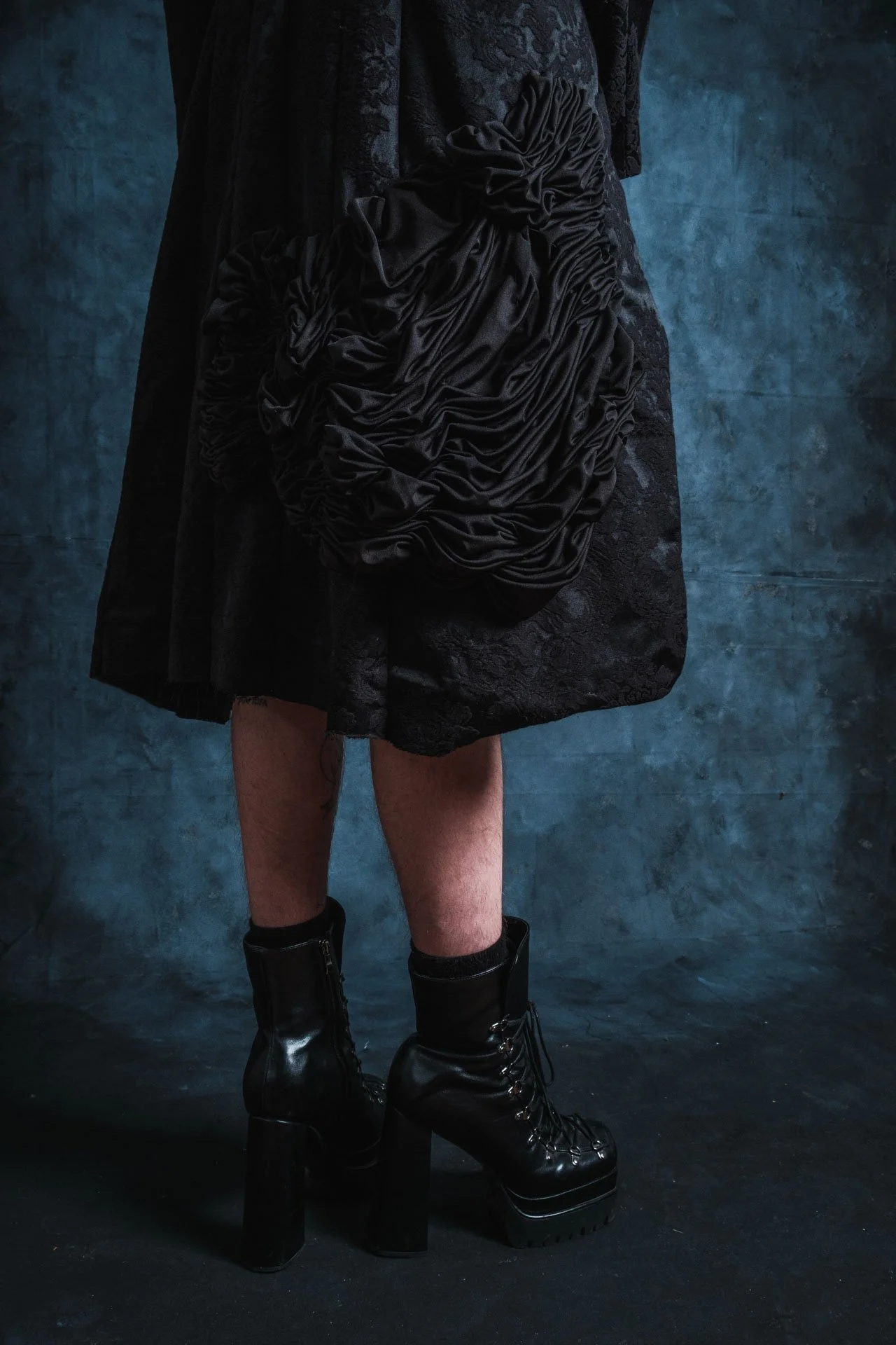 Une personne porte une robe noire avec des détails en tissu noué, des bottes à talons épaisses et une chaussette. La photo est prise contre un mur bleu foncé, mettant en valeur les détails de la tenue.