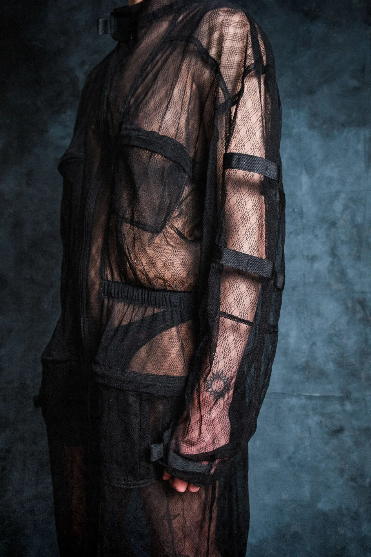 Une personne porte une robe noire en dentelle transparente, avec des motifs et des ceintures, contre un fond sombre.