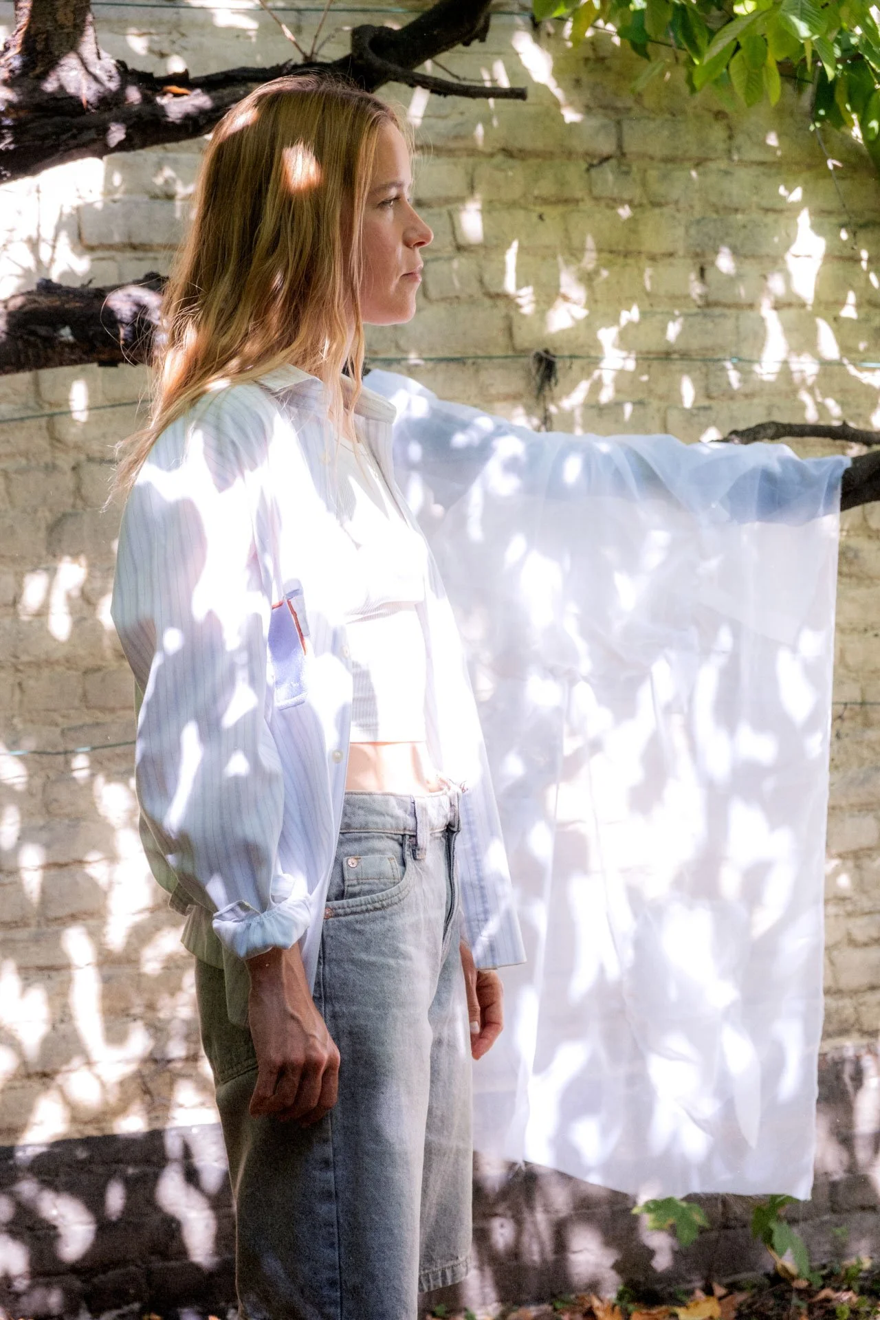 Femme debout en profil, portant une chemise blanche rayée et un jean, dans un lieu en plein air avec des ombres de feuilles sur un mur en pierre.
