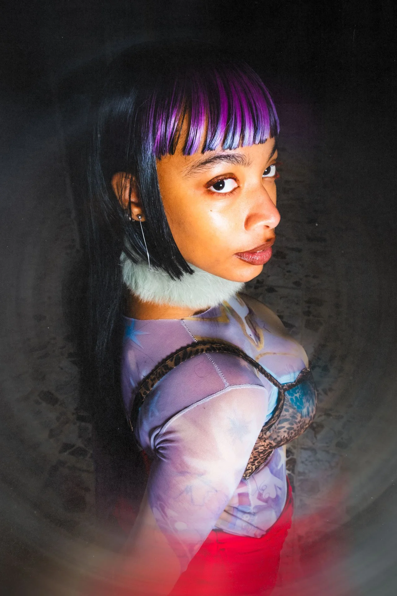Jeune femme portant une perruque noire avec des mèches violettes, une coiffure à frange, un haut coloré et une jupe rouge, regardant par-dessus l'épaule, dans une ambiance sombre.