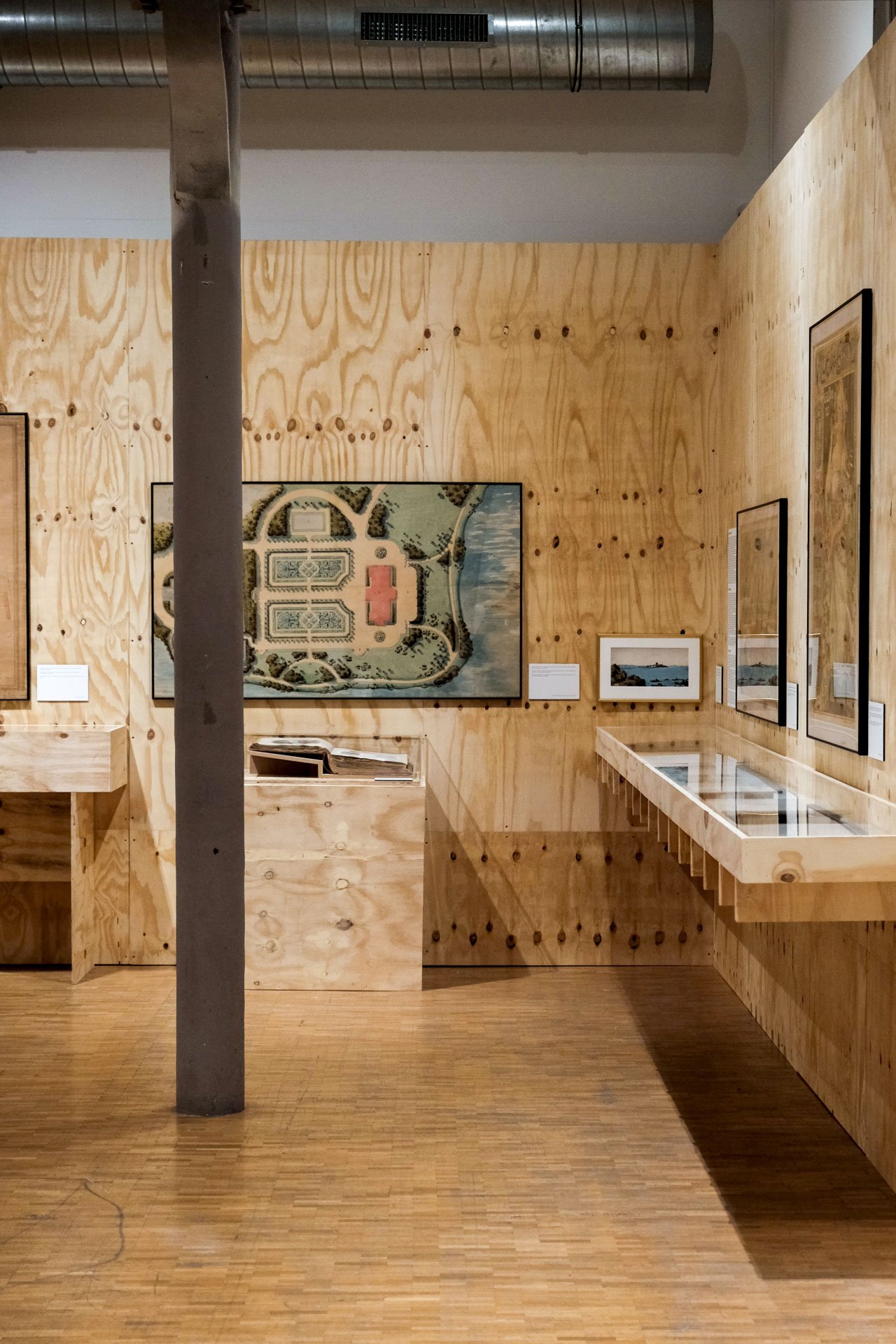 Une salle d'exposition avec des œuvres d'art encadrées accrochées à un mur en bois clair, avec des vitrines contenant des objets ou documents, et un sol en bois, un poteau en béton au centre.