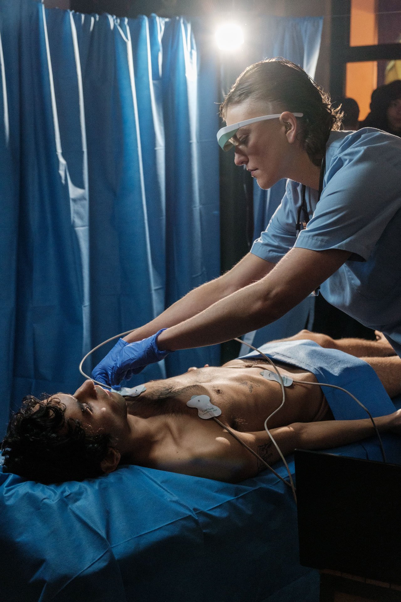 Un médecin effectue une intervention chirurgicale sur un patient couché sur une table d'opération. La médecin porte des lunettes de protection et des gants bleus, et travaille dans une salle d'hôpital avec des rideaux bleus.