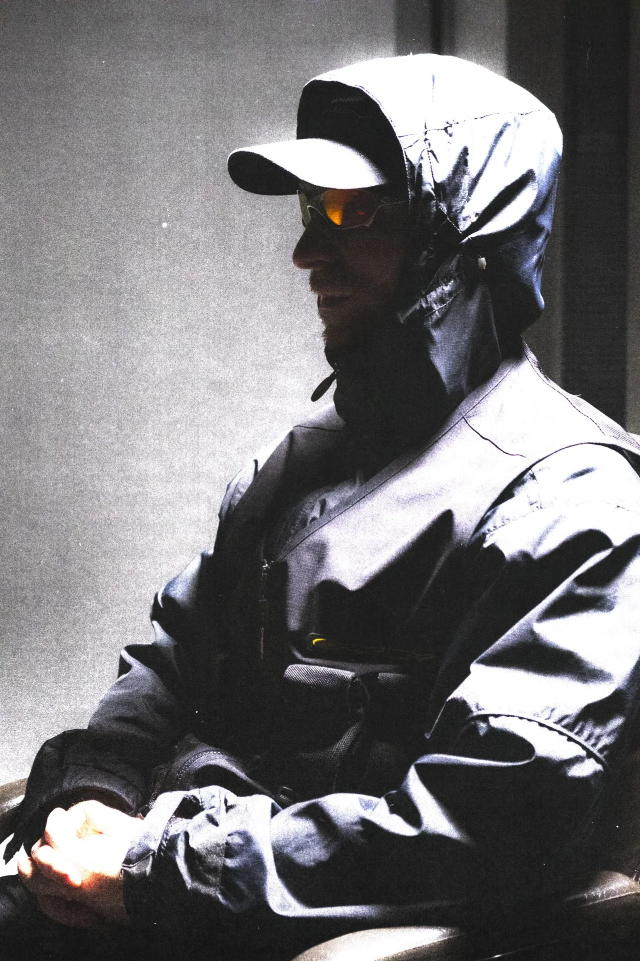 Un homme portant une casquette, des lunettes de soleil orange, une veste noire et blanche, et des gants noirs, semble assis ou debout dans un environnement sombre.