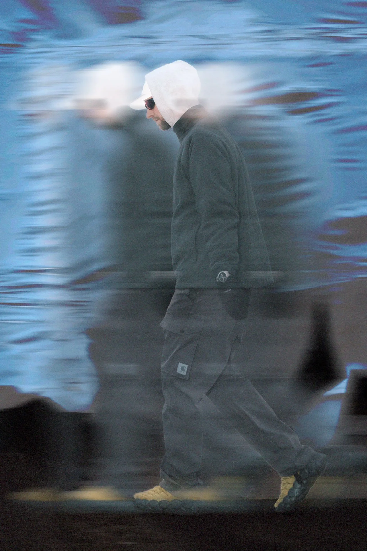 Un homme marche avec des vêtements décontractés, portant un sweat à capuche blanc et des lunettes de soleil, dans un environnement flou avec des couleurs bleues et grises.