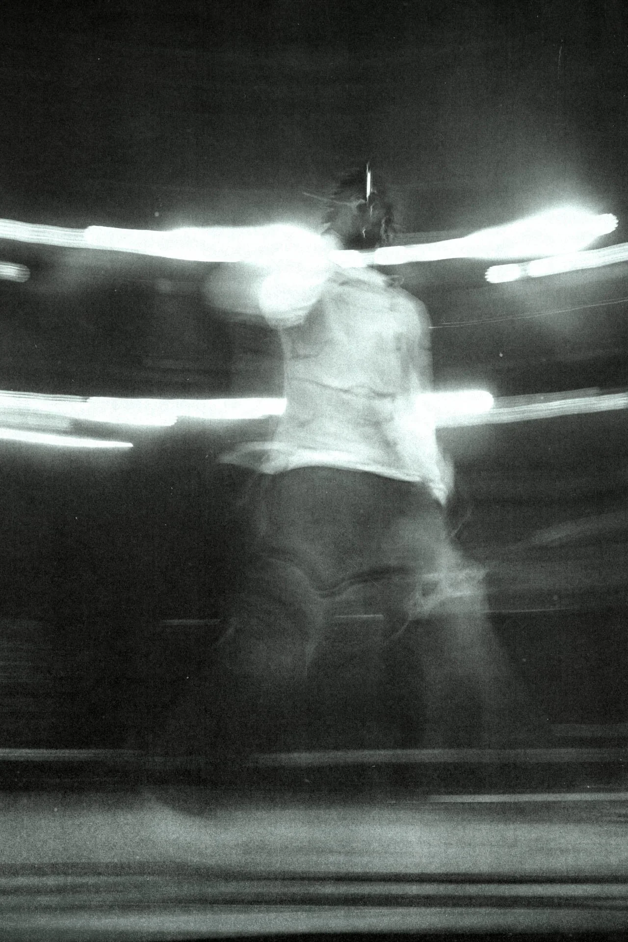 Photo floue d'une personne en mouvement, prise dans l'obscurité avec des traînées lumineuses. La personne semble porter un vêtement clair et se trouve devant des lumières. L'image est en noir et blanc.