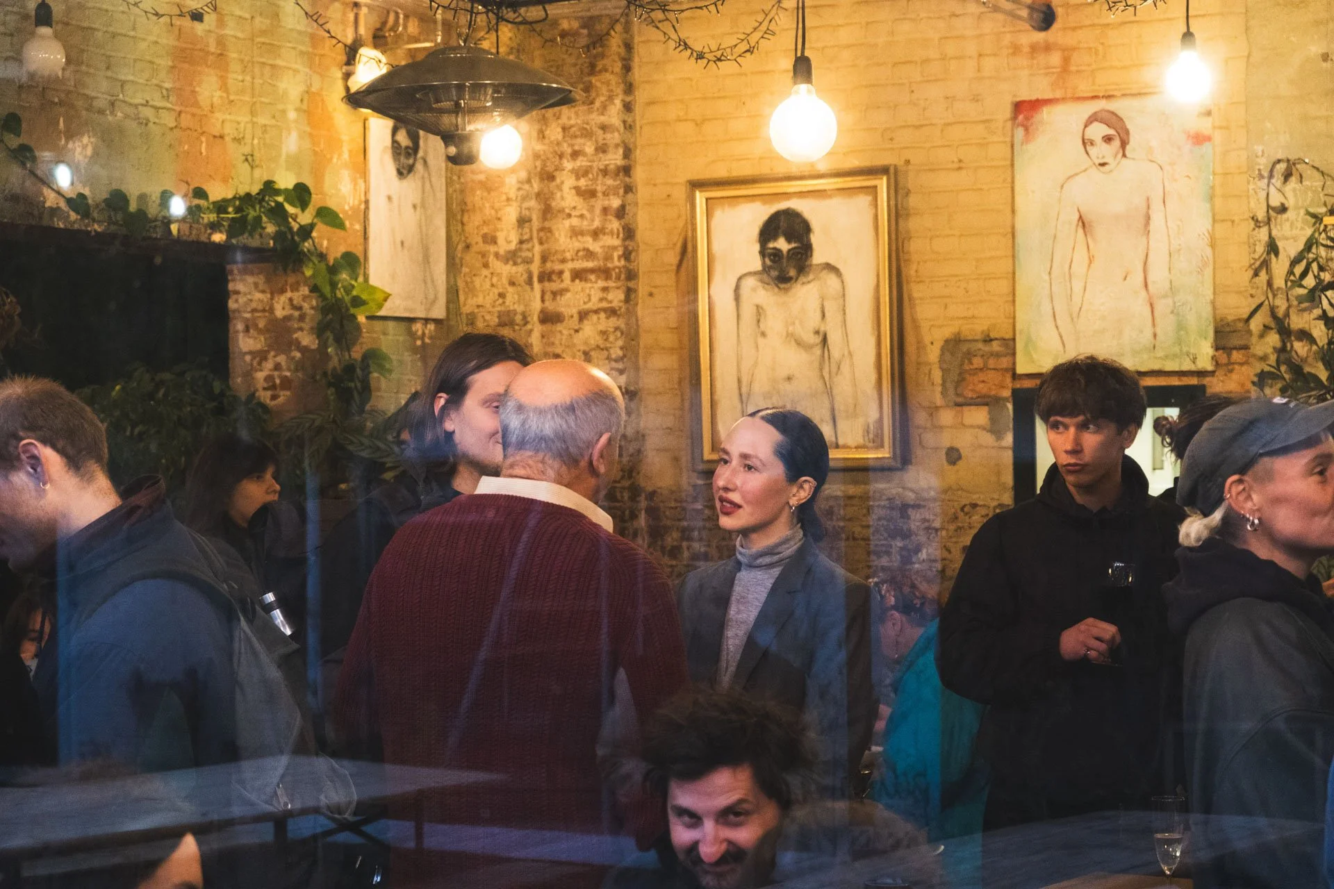 Une scène dans un café ou une galerie d'art où plusieurs personnes discutent, avec des œuvres d'art peintes de nus accrochées au mur en arrière-plan. L'ambiance est chaleureuse avec un éclairage tamisé.