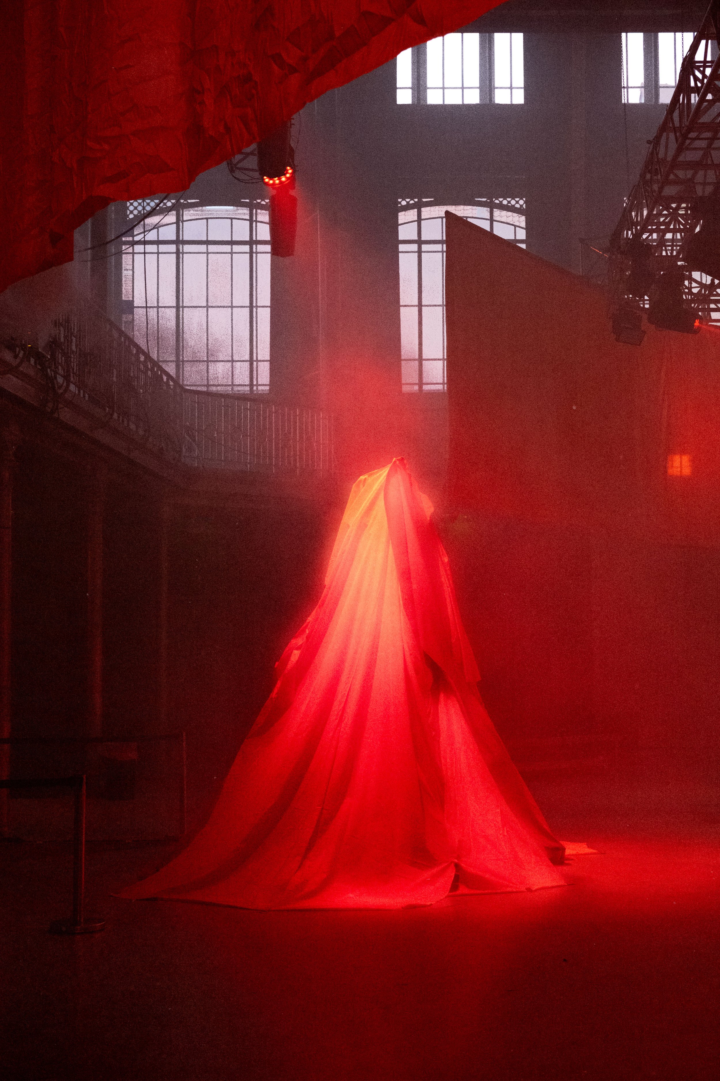 Une installation artistique dans une grande salle avec des tissus rouges suspendus et une lumière rouge qui illumine le sol. Des fenêtres en arrière-plan laissent entrer de la lumière naturelle