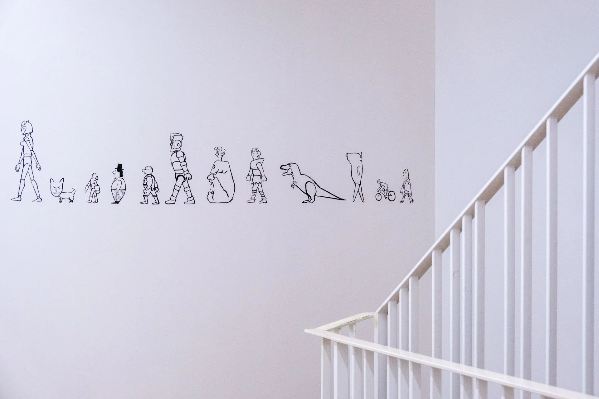 Décoration murale représentant l'évolution humaine et technologique avec des dessins en ligne noirs d'une variété de personnages de différentes tailles, formes et espèces, alignés horizontalement sur un mur blanc à côté d'une rampe d'escalier blanche