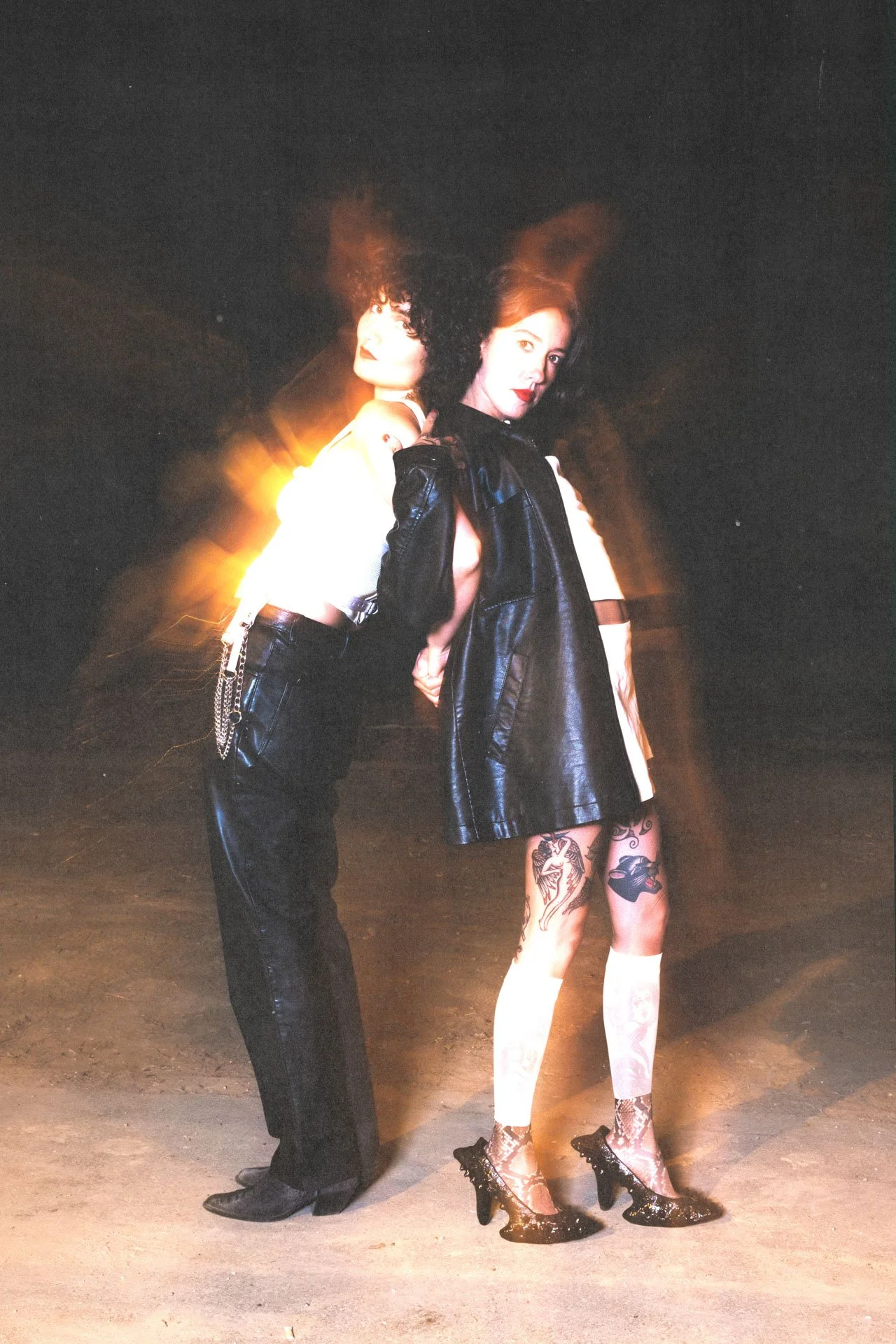 Deux femmes posant devant un fond sombre. La femme à gauche a des cheveux bouclés, porte une veste noire, un pantalon noir avec une chaîne, et des chaussures noires. La femme à droite a des cheveux longs, portent une veste en cuir noire, une jupe ou 