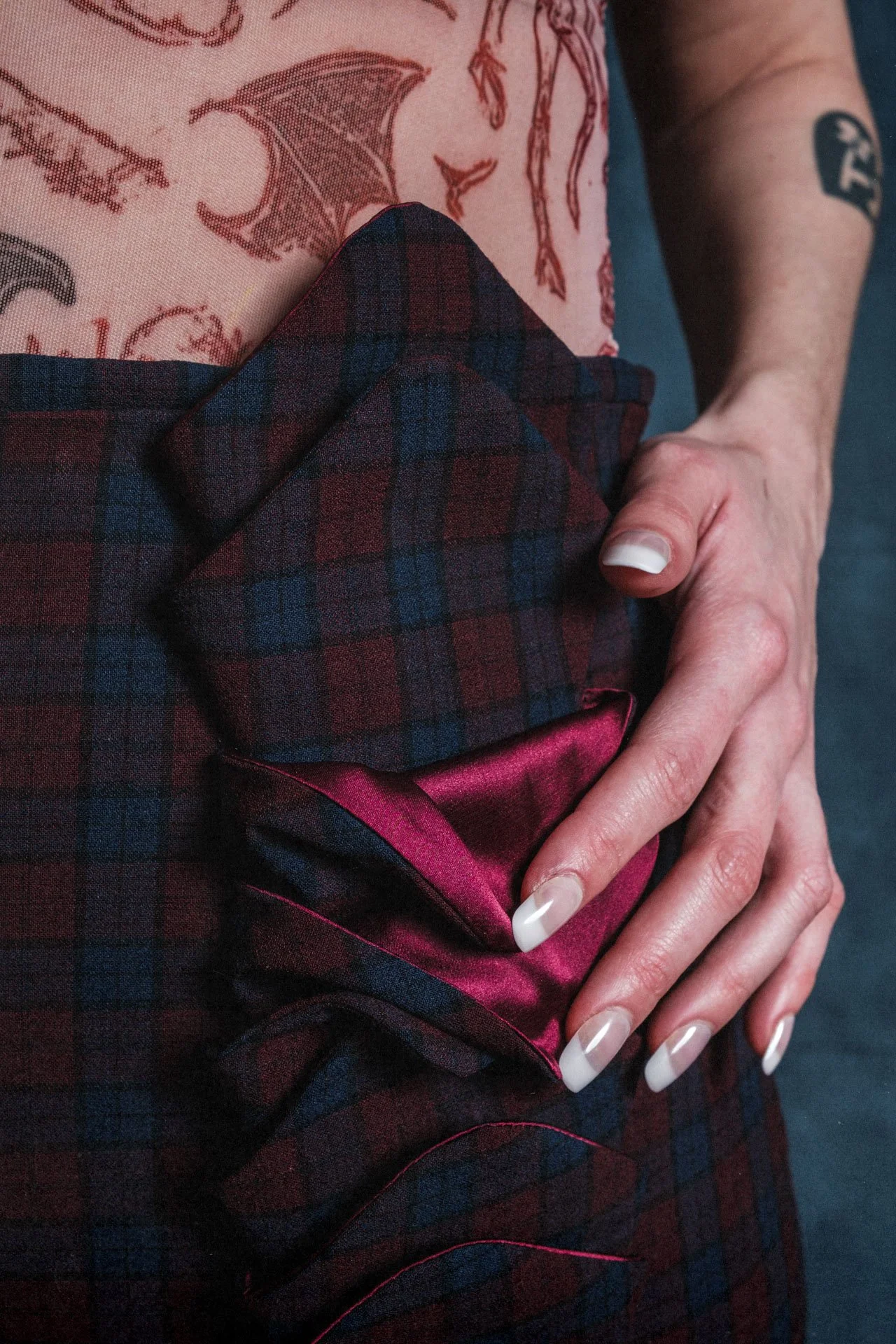 Une main délicate repose sur une robe en tartan rouge, vert et bleu, avec une doublure en satin rose brillant. La personne porte un tatouage de dragon sur le bras.