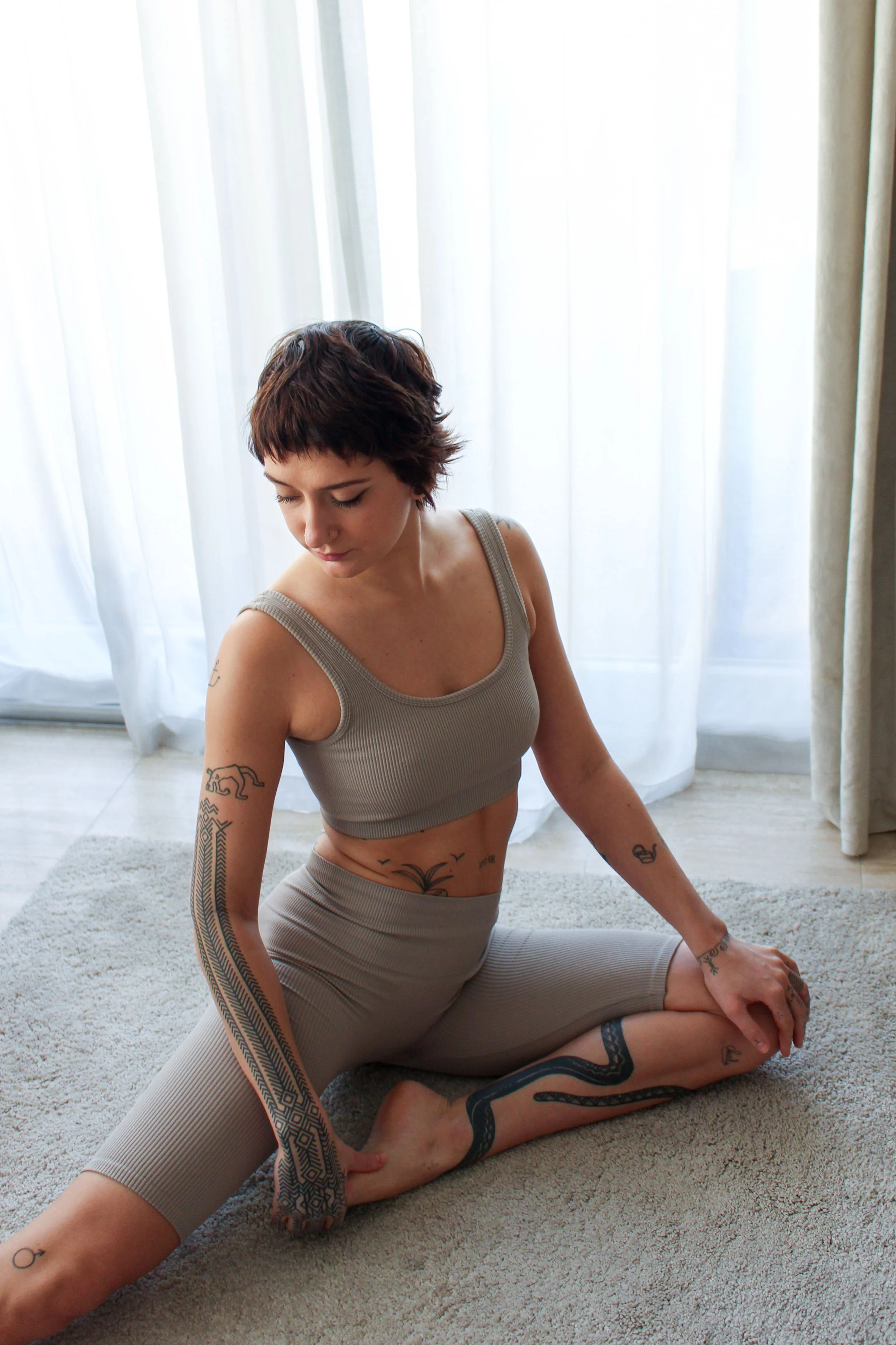 Une femme aux cheveux courts et bruns, portant un débardeur beige et un legging beige à motifs, pratiquant le yoga sur un tapis en intérieur avec des rideaux blancs en arrière-plan.