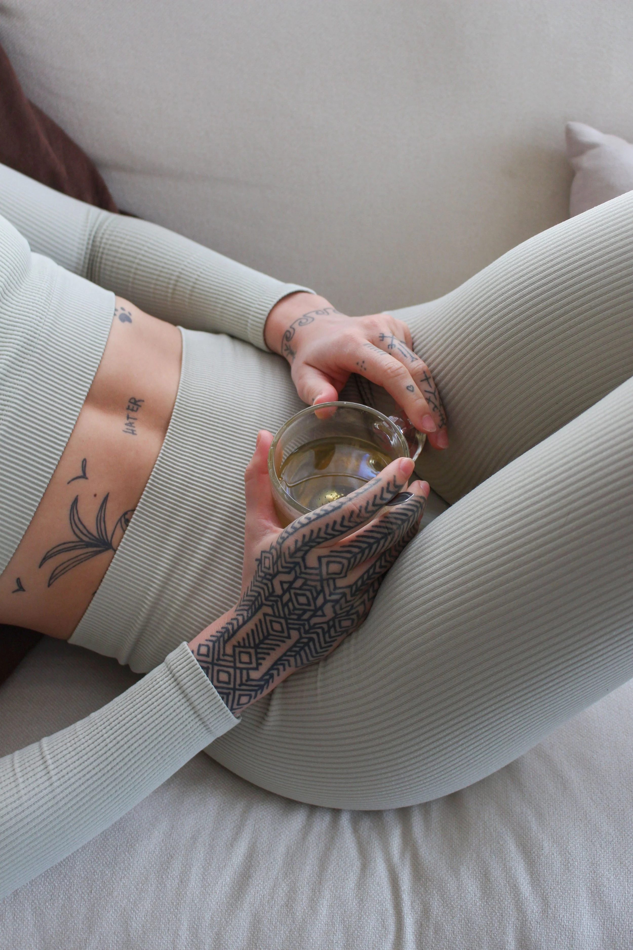 Personne assise sur un lit, portant des vêtements en maille beige, tenant un verre d'eau. La personne a des tatouages sur une main et l'autre bras, et une fois sur le ventre avec le nom "LATER" écrit sur le côté.