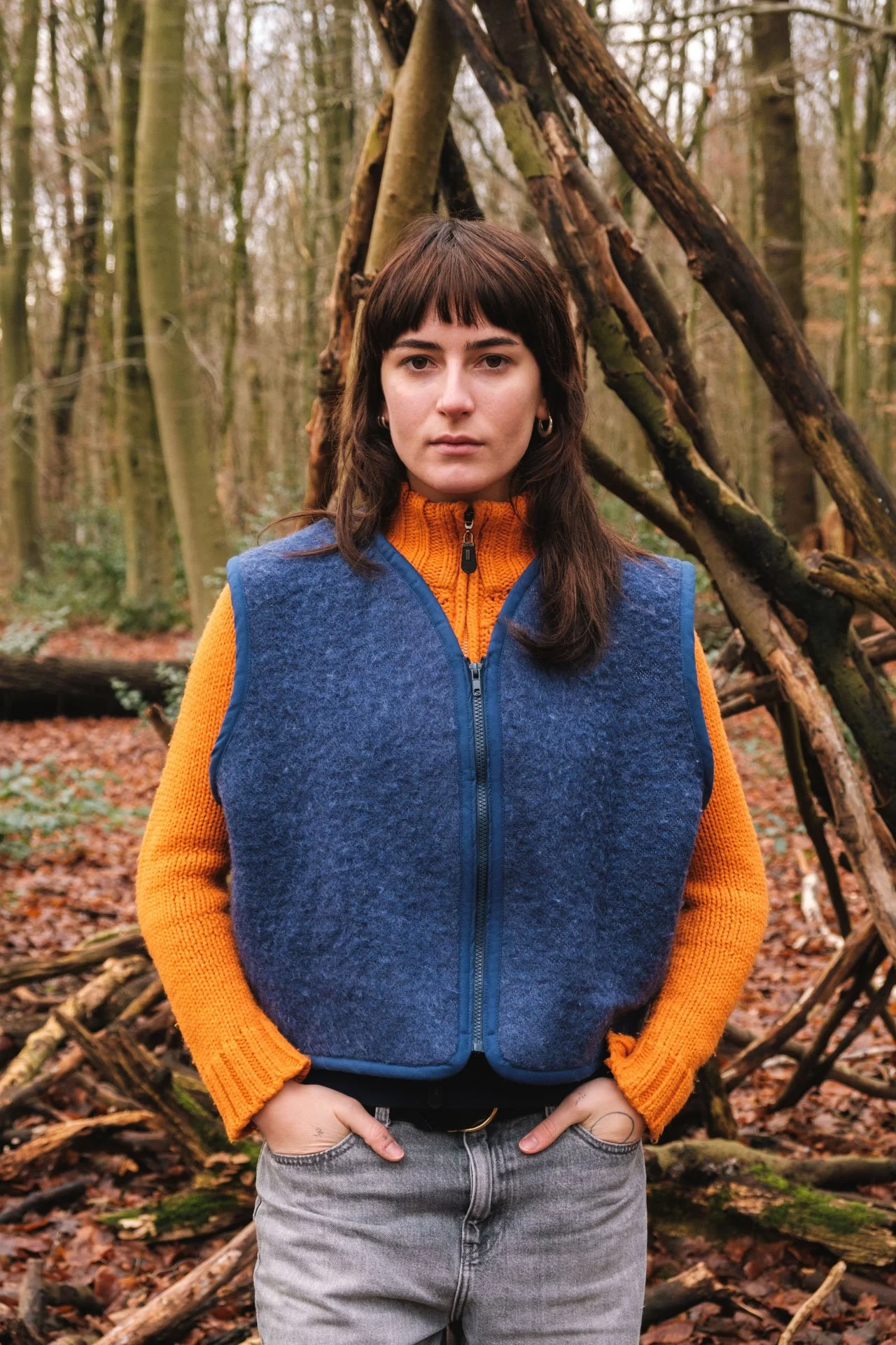 Une femme debout dans une forêt, portant une veste à zip bleu marine avec un intérieur orange, et un pull orange en dessous. Elle a les cheveux bruns avec une frange et des boucles, et regarde directement l'appareil photo.